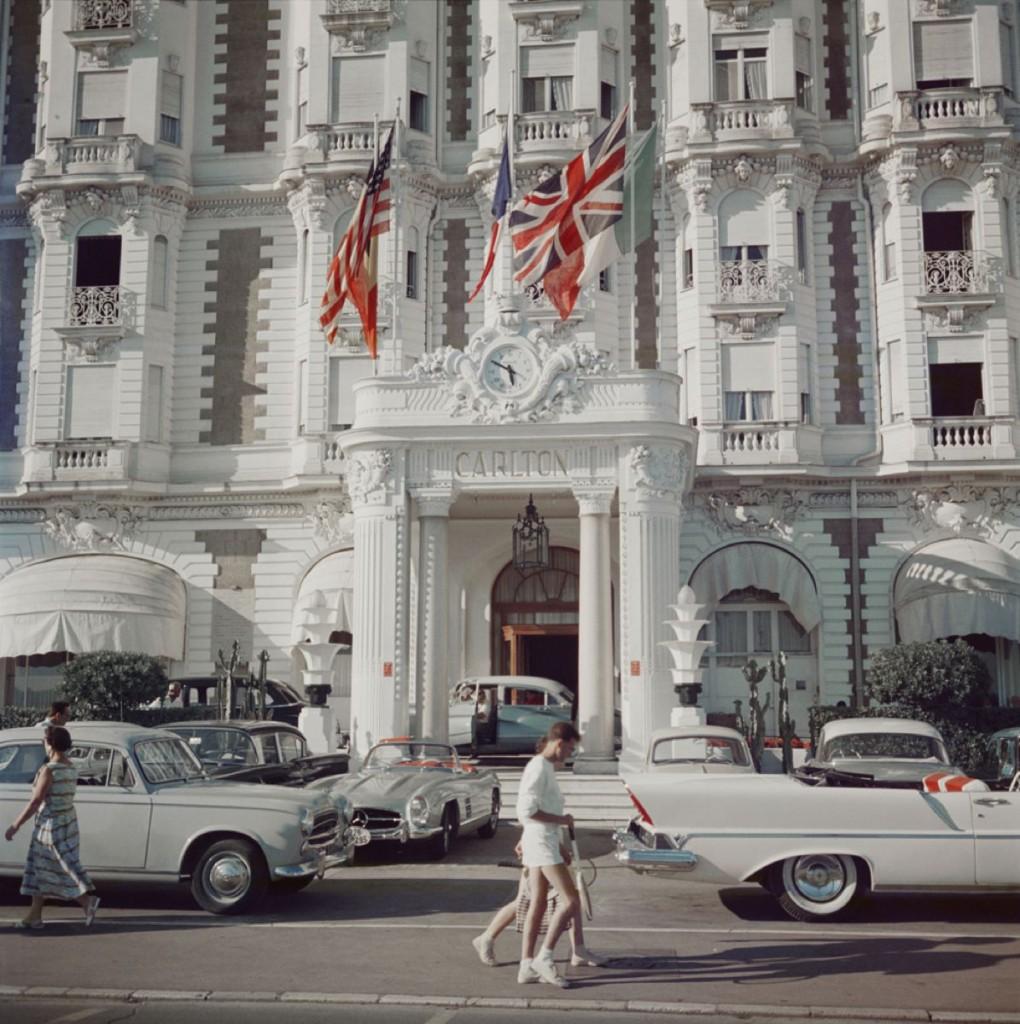Carlton Hotel" von Slim Aarons

Der Eingang zum Carlton Hotel, Cannes, Frankreich, 1958.

Ein elegant gekleidetes Paar in weißer Tenniskleidung mit Schlägern in der Hand schlendert am Eingang des kultigen Carlton Hotels vorbei. Vor dem Hotel parken