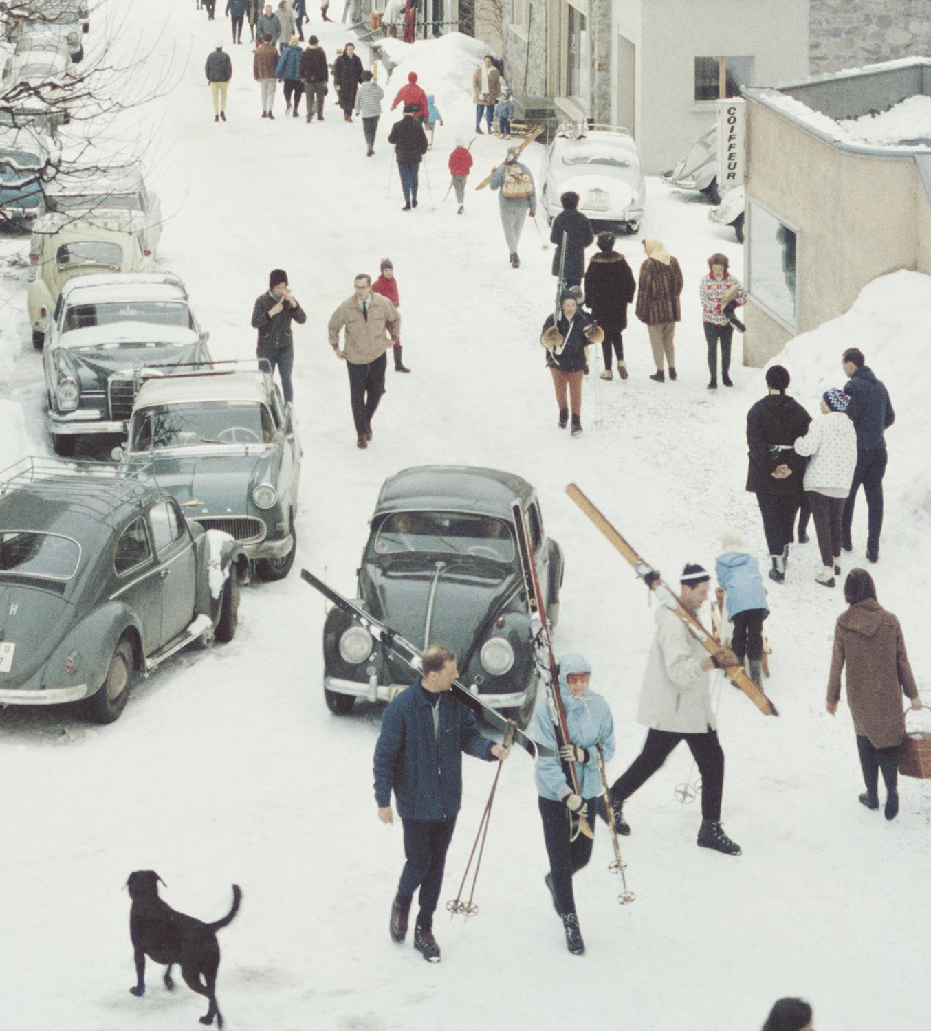 Klosters 1963 Slim Aarons

Skiers pass by the Hotel Chesa Grischuna in Klosters, 1963.

Skiers walk past the hotel and along the snow covered road carrying skis on their shoulders. The road is lined with classic and vintage cars of the era and