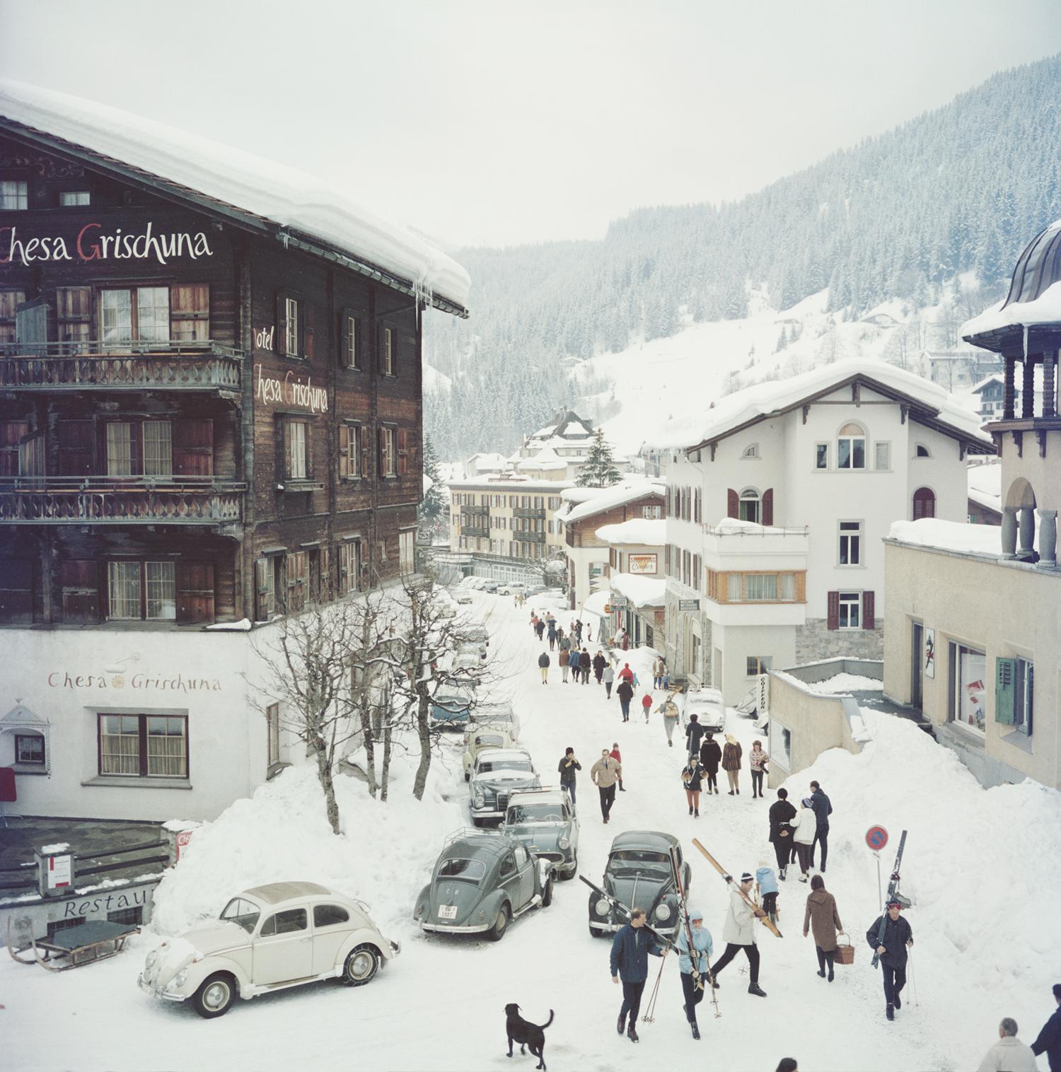 Klosters by Slim Aarons

Skiers pass by the Hotel Chesa Grischuna in Klosters, 1963.

Skiers walk past the hotel and along the snow covered road carrying skis on their shoulders. The road is lined with classic and vintage cars of the era and