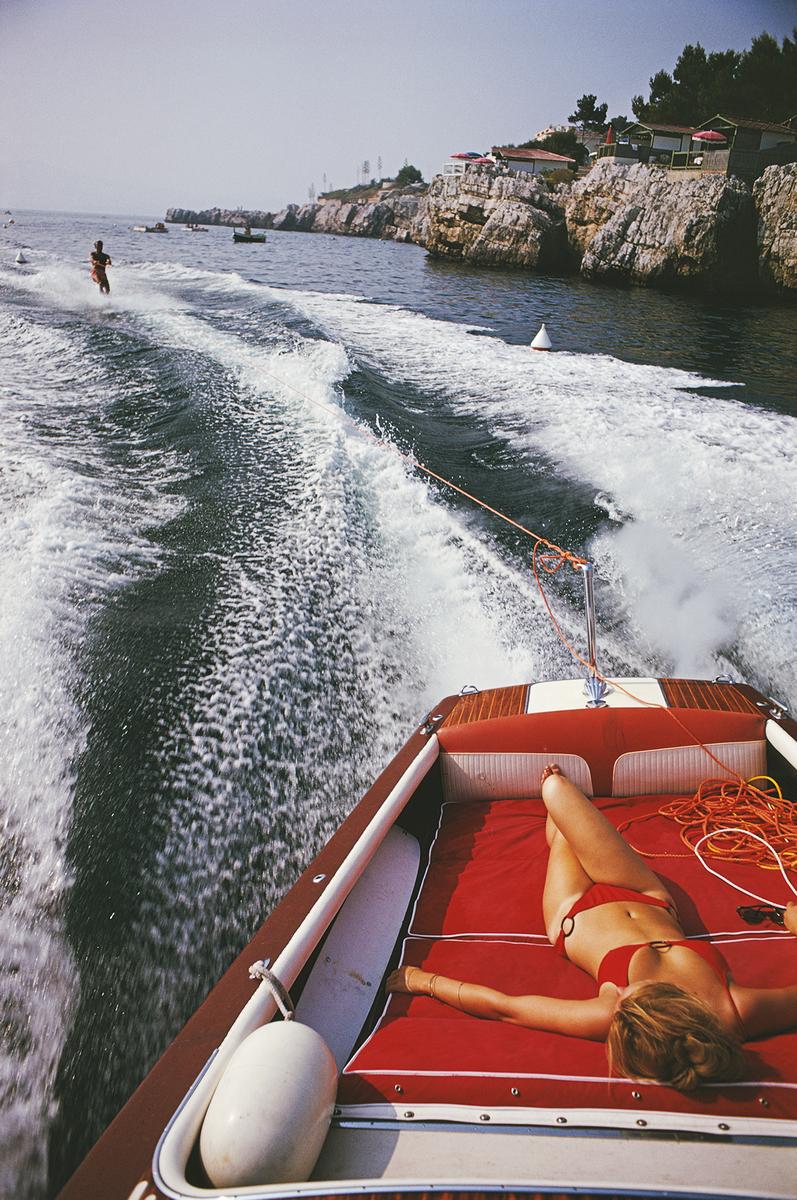 Slim Aarons Nachlass-Druck  - Freizeit in Antibes 1969

Eine Frau sonnt sich in einem Motorboot, das einen Wasserskifahrer zieht, auf dem Meer vor dem Hotel du Cap-Eden-Roc in Antibes an der französischen Riviera, August 1969. (Foto: Slim