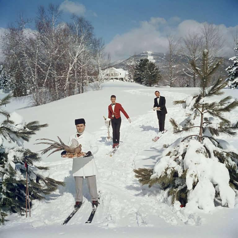 Drei Skikellner auf einer Skipiste, wobei der Mann im Vordergrund einen Vogel auf einem Tablett trägt, der zweite Mann einen Wein in einem Eiskübel und der dritte eine Speisekarte, um 1960.

Slim Aarons (1916-2006), der zunehmend für seinen Einfluss