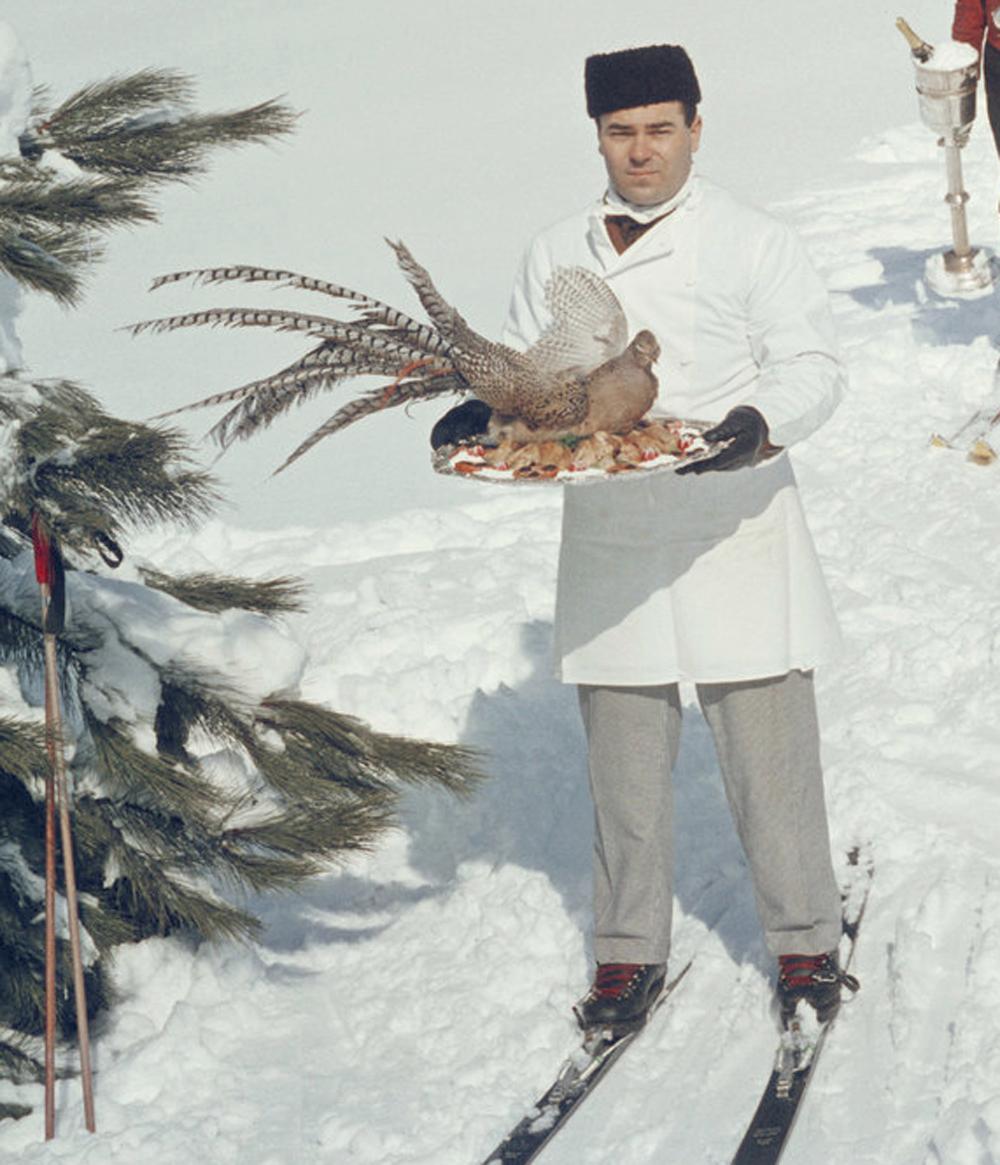 Three skiing waiters on a ski slope, circa 1960. In the foreground, a white-clad chef skis towards the viewer, offering a beautifully prepared bird on a tray. Behind him, a man in a formal maroon red jacket carries fine wine in a silver ice bucket.