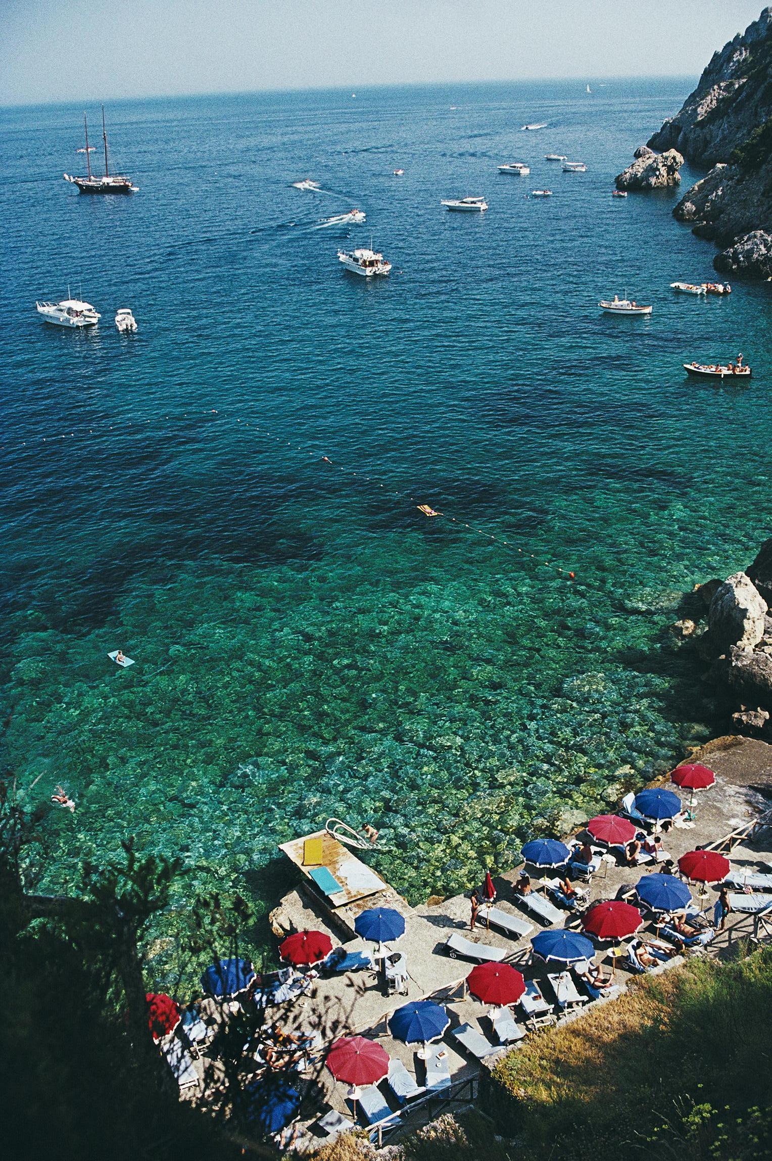 Aerial view with sunbathers and parasols and, dotted with yachts and small boats, the waters off the coast of Porto Ercole, Italy, in July 1991. The image was taken from Hotel Il Pellicano. 

Slim Aarons
View From Il Pellicano
1991
C print (Printed