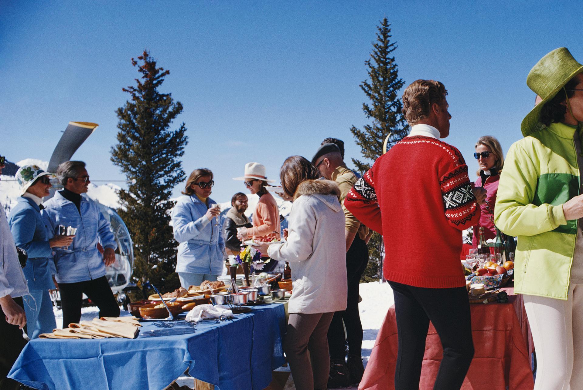 Cette photographie de la fin des années 1960, prise par le photographe mondain Slim Aarons, montre deux femmes portant des vêtements de ski aux couleurs vives, au premier plan d'un groupe de personnes participant à une fête à Snowmass Village, dans