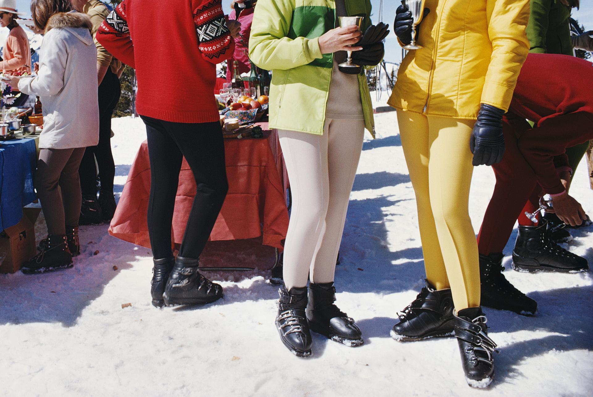 Questa foto ritratto della fine degli anni '60, scattata dal fotografo di società Slim Aarons, ritrae due donne, che indossano abiti da sci dai colori vivaci, in primo piano rispetto a un gruppo di persone che partecipano a una festa a Snowmass