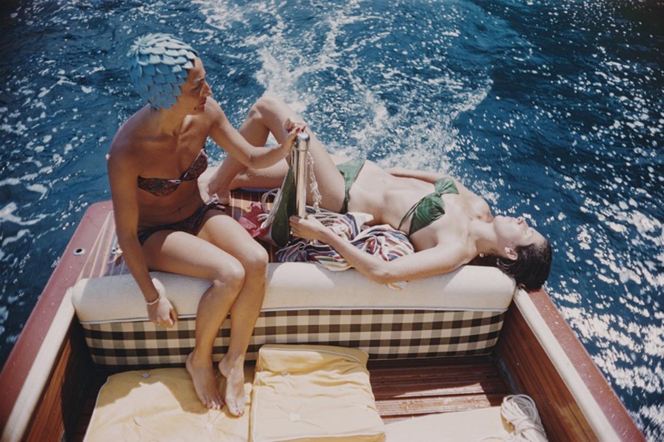 Vuccino And Rava 
1958
by Slim Aarons

Slim Aarons Limited Estate Edition

Carla Vuccino, wearing a swimming cap, and a sunbathing Marina Rava, both wearing bikinis as they sit on the rear of a boat, on the waters off the coast of the island of