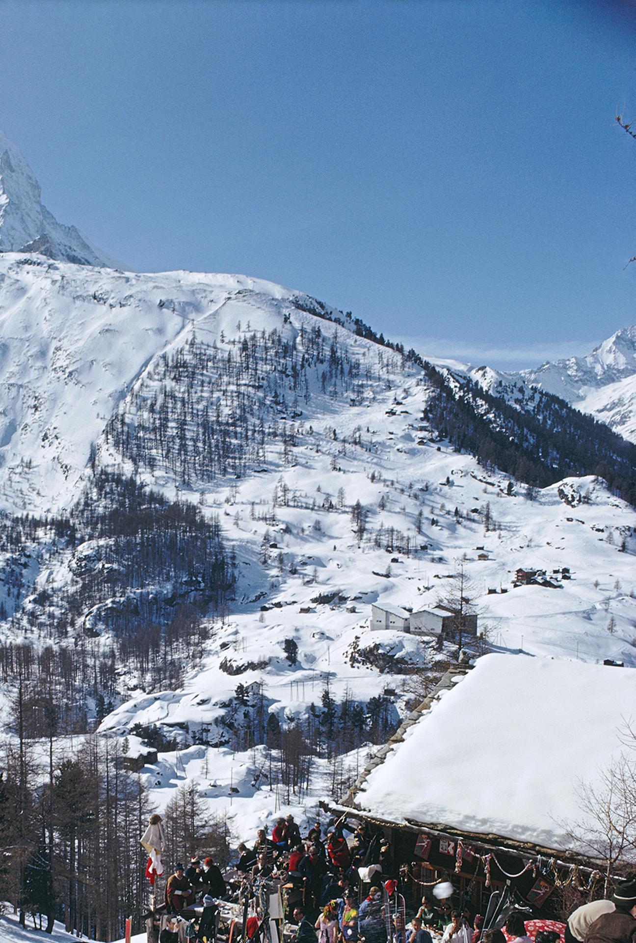 Diese Landschaftsfotografie aus der Mitte des Jahrhunderts, aufgenommen vom Society-Fotografen Slim Aarons, zeigt das Après-Ski in Zermatt, Schweiz, im März 1968. 

In dieser Szene von lässigem Winterglamour erheben sich verschneite Berge vor einem
