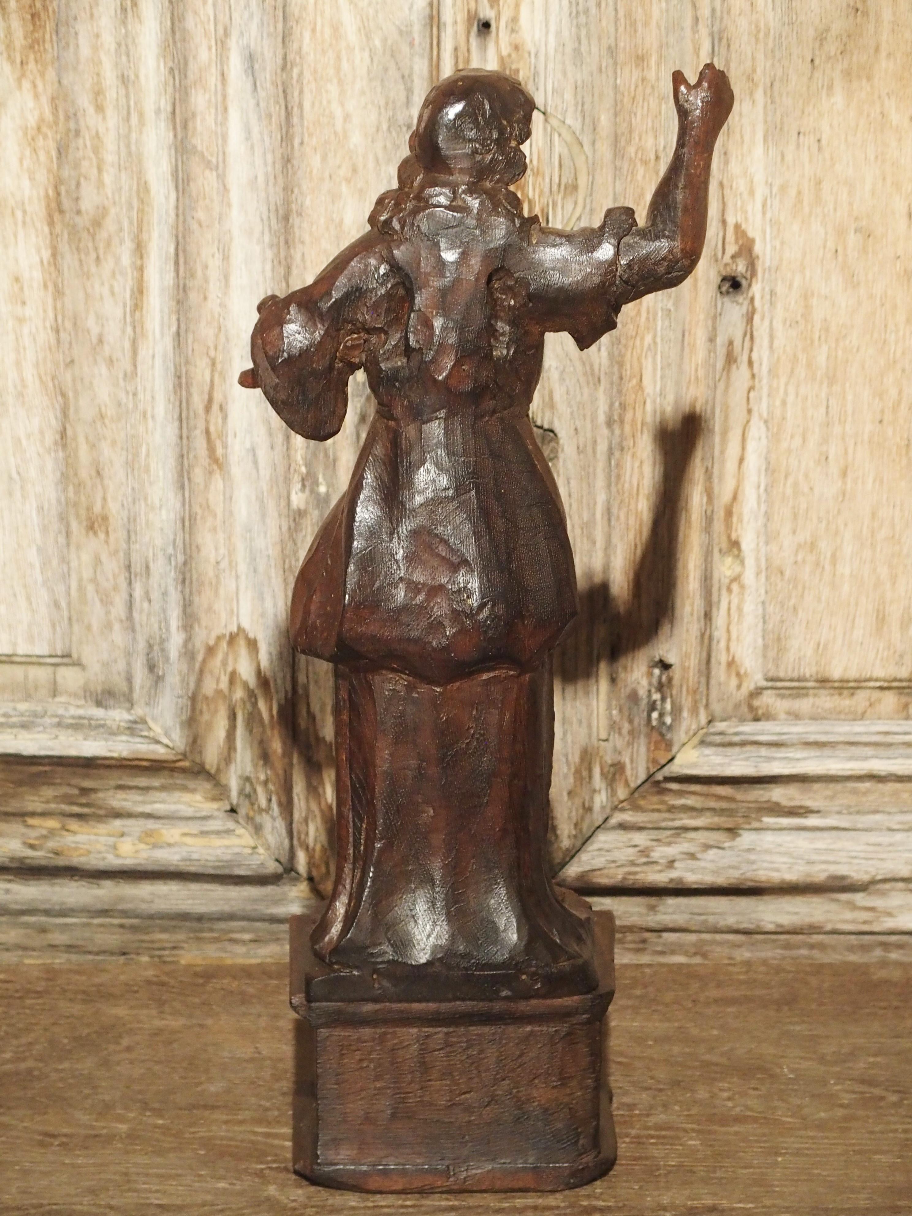 This small French statue from the 1600s depicts a person in a two- piece robe with the bottom right hand portion of the robe attached above the knee. He is standing on a small carved pedestal with carved escutcheon motifs surrounding a date of 1520.