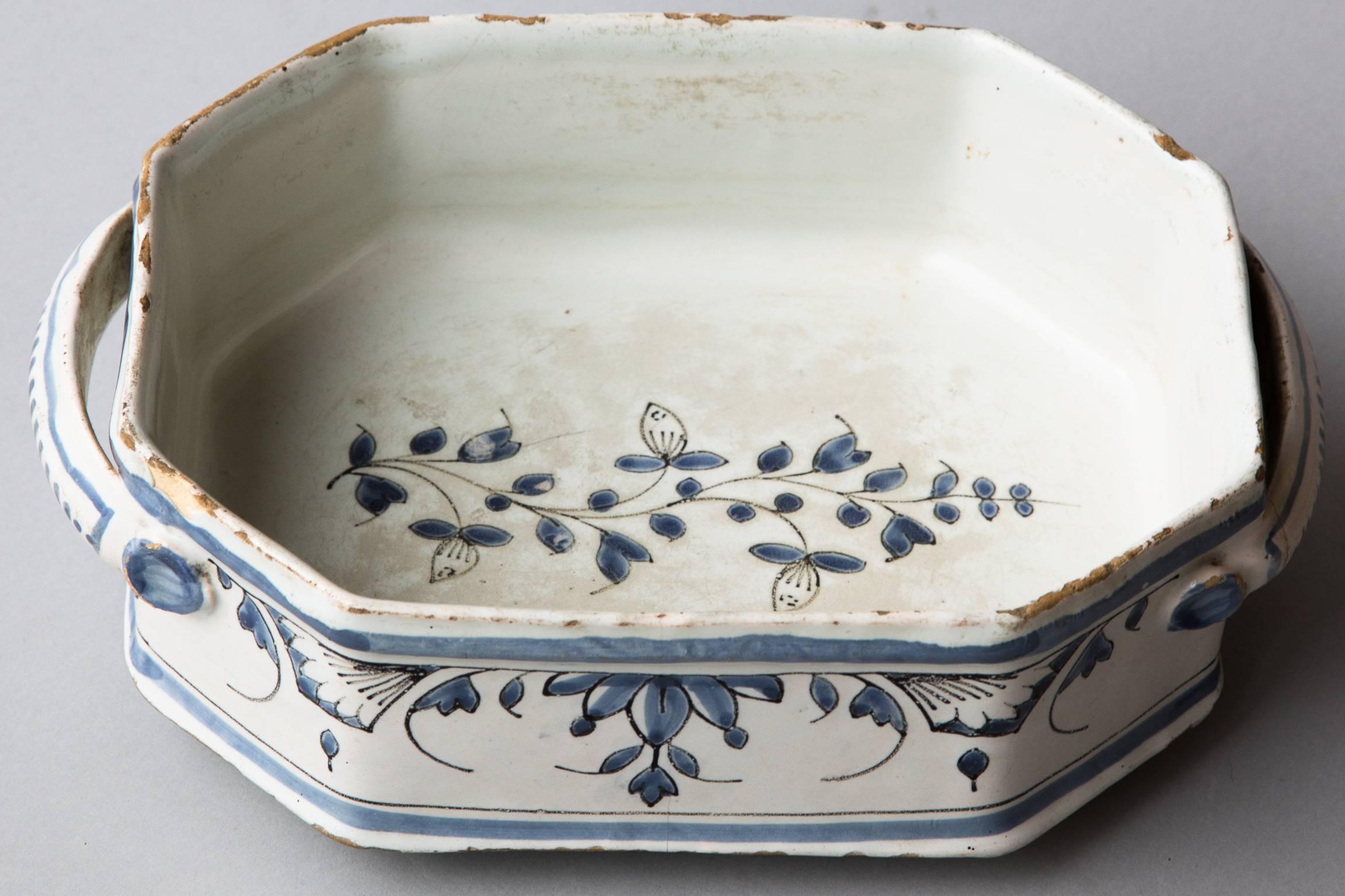 The octagonal body standing on three bun feet with two handles at either end. 
Decorated with geometric floral designs in blue and black on a milky white background. 
Rouen, France circa 1760. Old chips and scratches to the glaze and an old repair