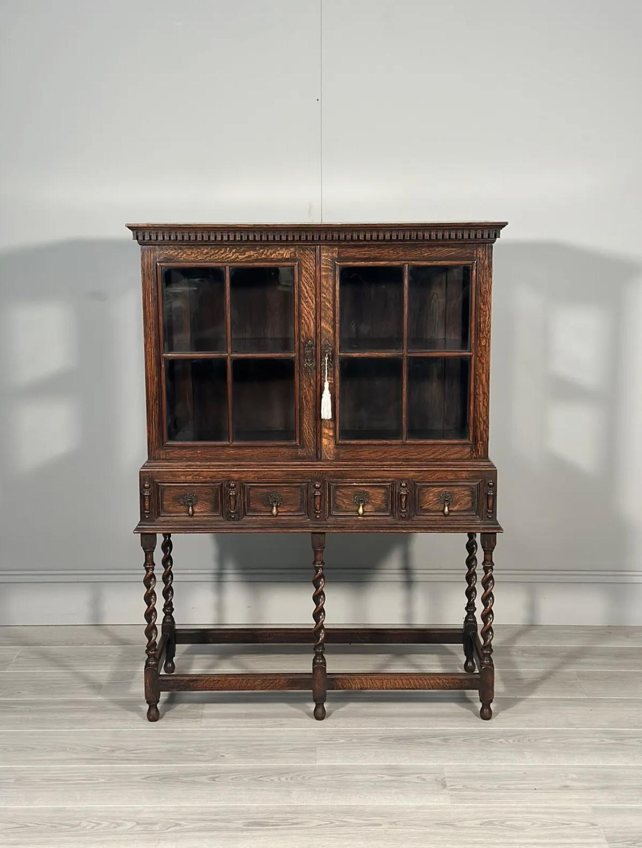 A small oak display cabinet dating to the 1920’s in a Jacobean style. The cabinet has front double doors with a working lock and key with internal shelf, glazed sides and sits on a 2 drawer base with barley twist legs, In overall very good