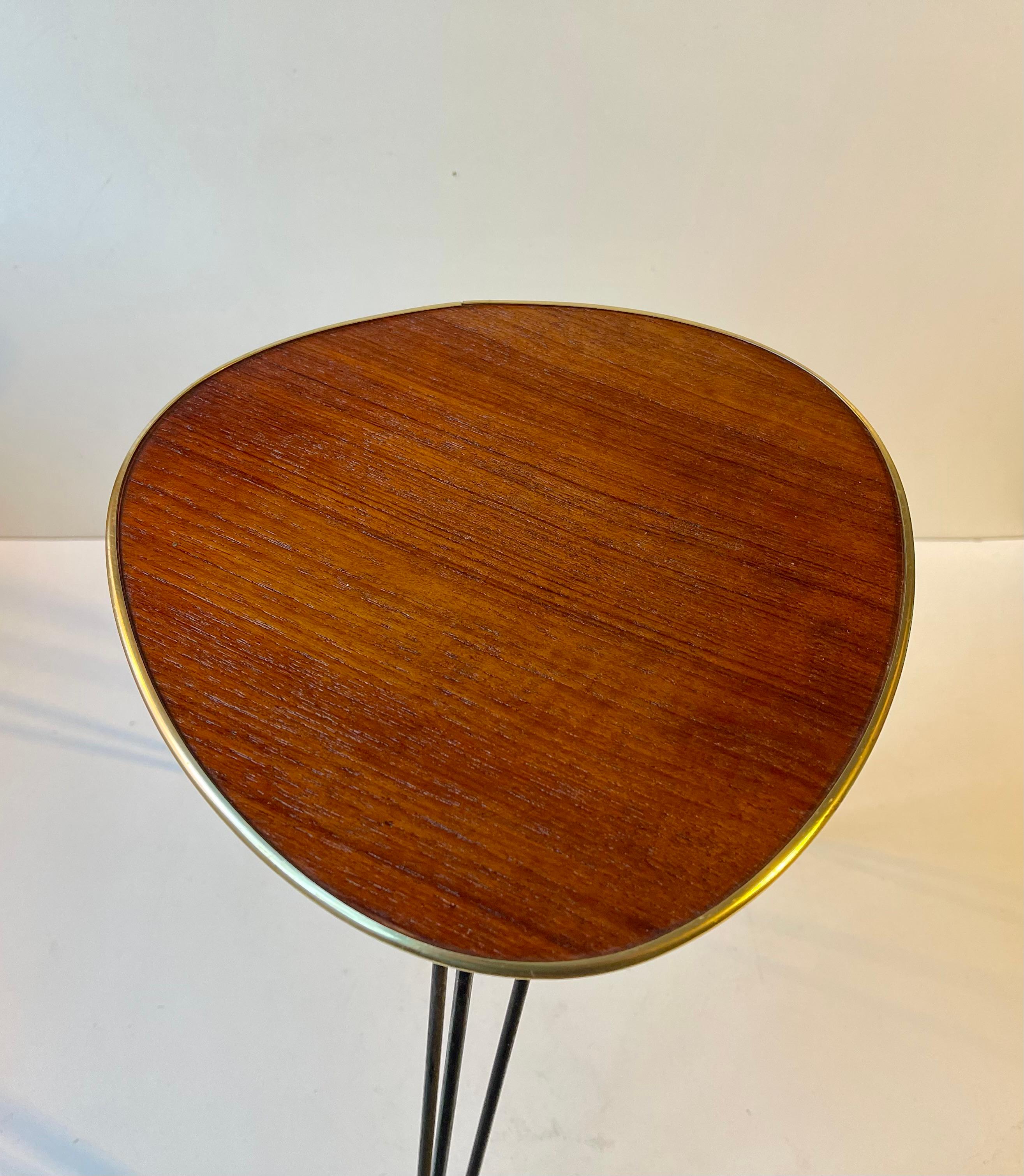 A small side, lamp or plant table featuring a guitar-pick shaped table top in teak with brass alloy accents and bed wired steel legs. A very graphic look due to its simple construction and 'light' appearance. Made in Scandinavian or Germany circa