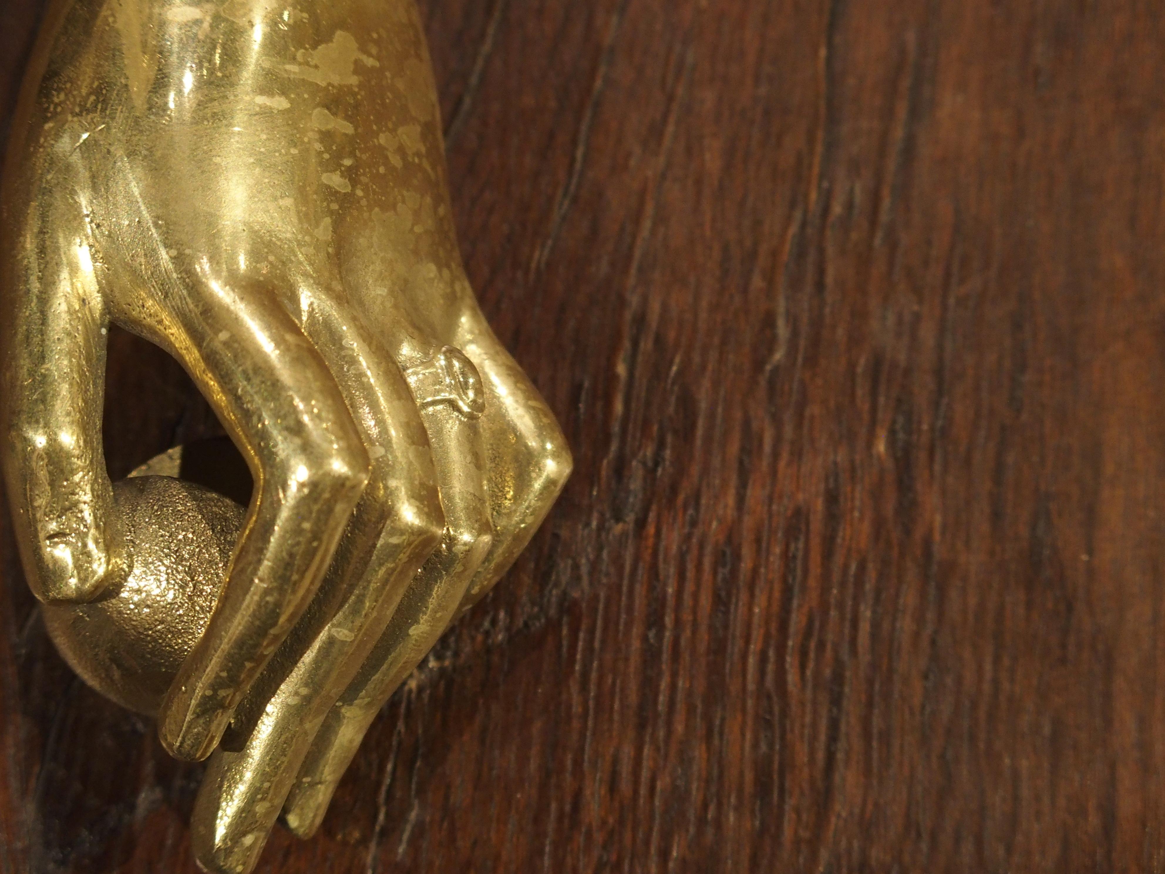 This small gilt bronze door knocker was crafted in France in the 1800s.

The door knocker depicts a left hand with slender fingers surrounding a small ball striker. The ring finger is adorned with an oval ring while the wrist is covered by an