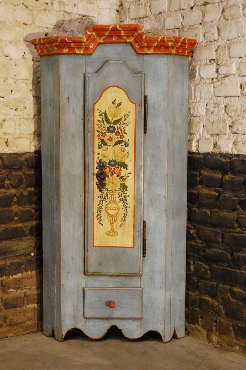 This small pine corner cabinet is made in in the early 1900s in Germany. It has a blue-gray base color and a handsomely decorated door. 
The country-style decoration depicts flowers and leaves in a vase below the door sits a small drawer with a