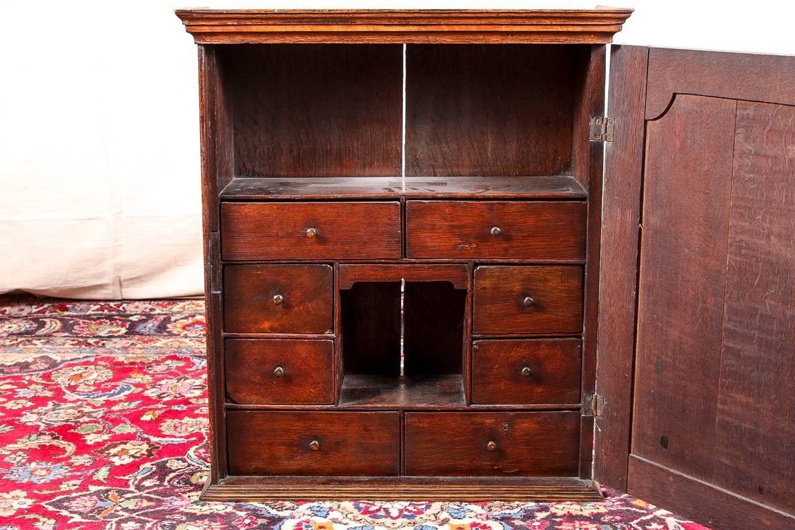 Small antique Georgian cabinet, oak, circa 1780, carved cornice and lower frame, the single door with recessed shaped panel framed in walnut, opening to large and small compartments and multiple drawers, with a brass escutcheon with key.