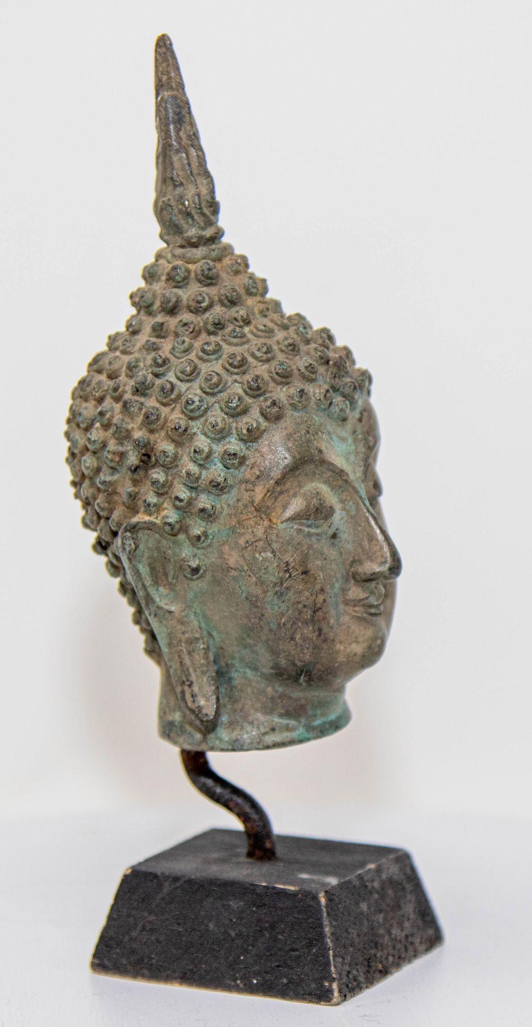Small antique Southeast Asian cast bronze buddha head sculpture with verdigris patina mounted on a black base wood stand.
Bronze Sukhothai style Buddha bronze head with green patina, from North Thailand.
The head is sensitively rendered with a