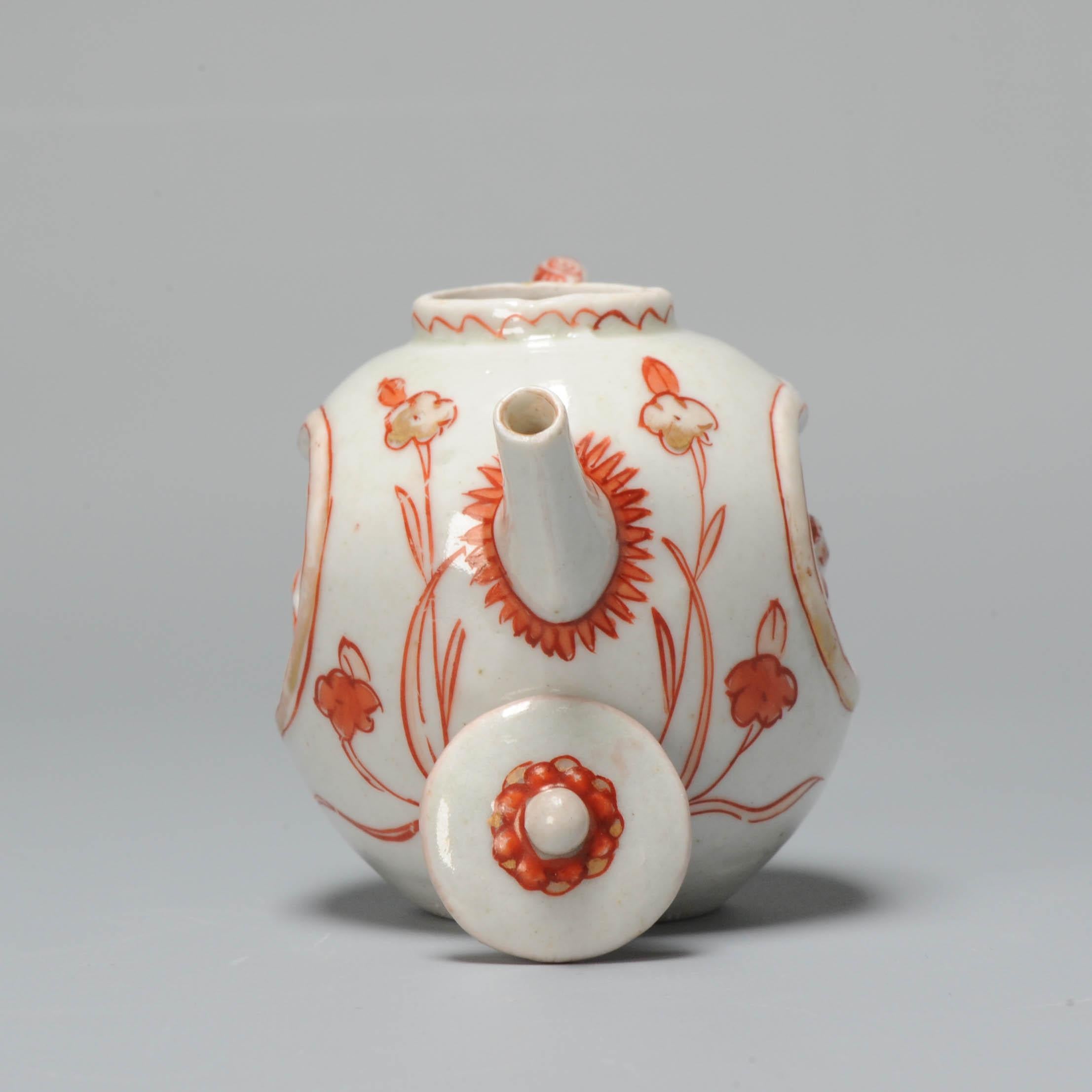 Small teapot, square, rounded body on flat unglazed base. Curved handle and a short straight spout. Small upright mouthrim, flat lid with round knob. Imari decorated in iron-red and gold on the sides with flowering plants and grasses and two deeply