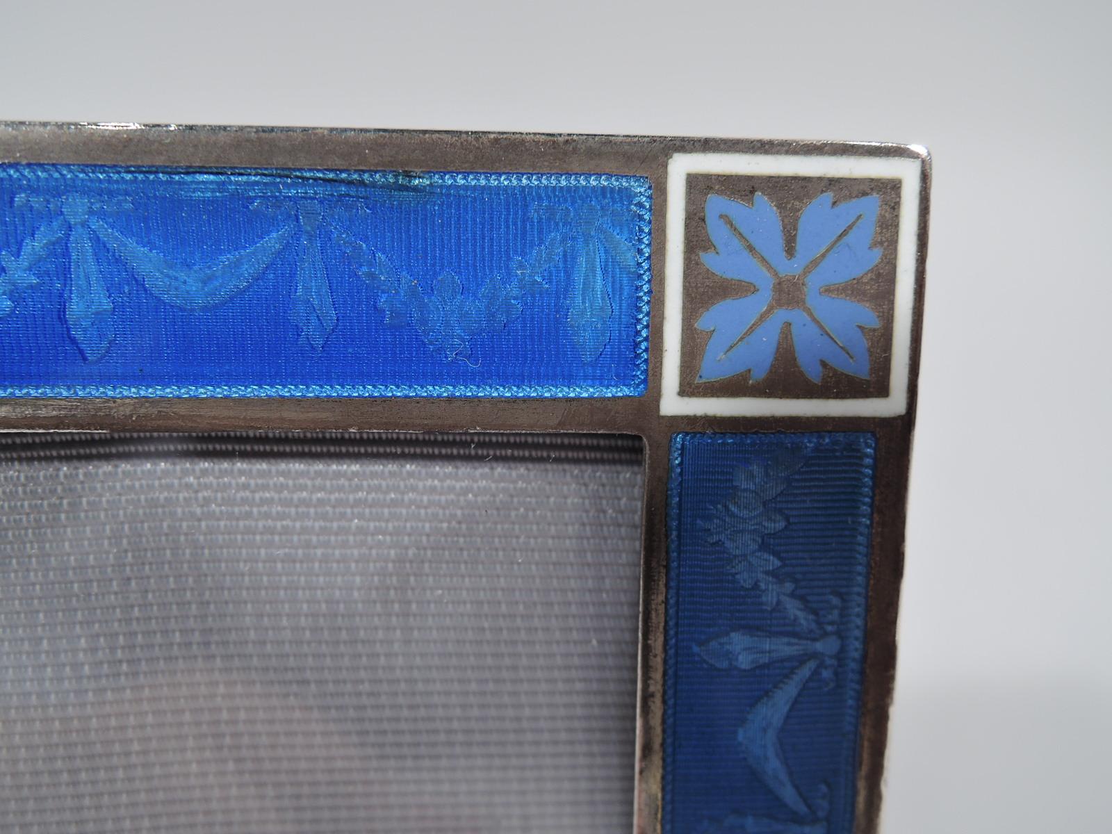 Edwardian Regency silver and enamel picture frame, ca 1910. Rectangular window with flat surround. Bow-tied floral garland on blue lined ground. Corner squares have white borders and are inset with violet paterae. With glass, silk lining, and velvet