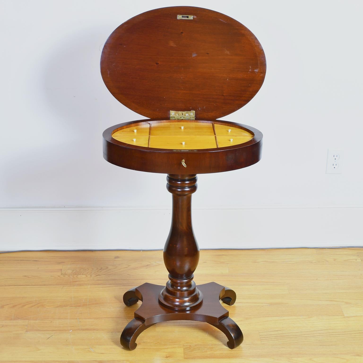 A handsome early 20th century small salon table in a dark cocoa-colored mahogany with oval top on turned pedestal column with quatre-form base resting on scrolled feet. Hinged-top lifts to access a light-wood interior (likely birch) with six lidded