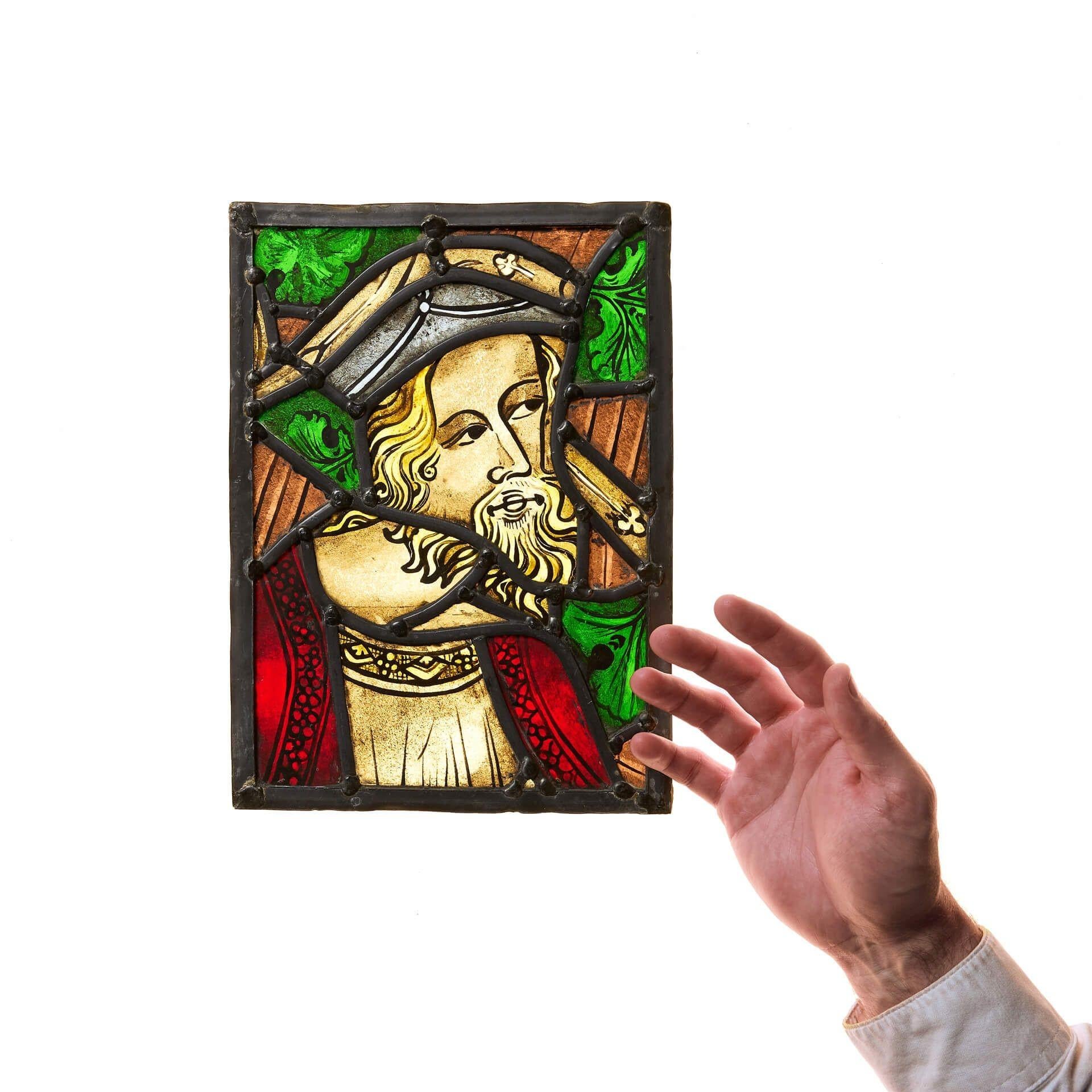 A small and well-captured antique stained glass window panel depicting a nobleman with blonde hair and bushy beard, dressed in red and wearing a cap. Though this piece dates from the early 20th century, the styling is older, reminiscent of medieval