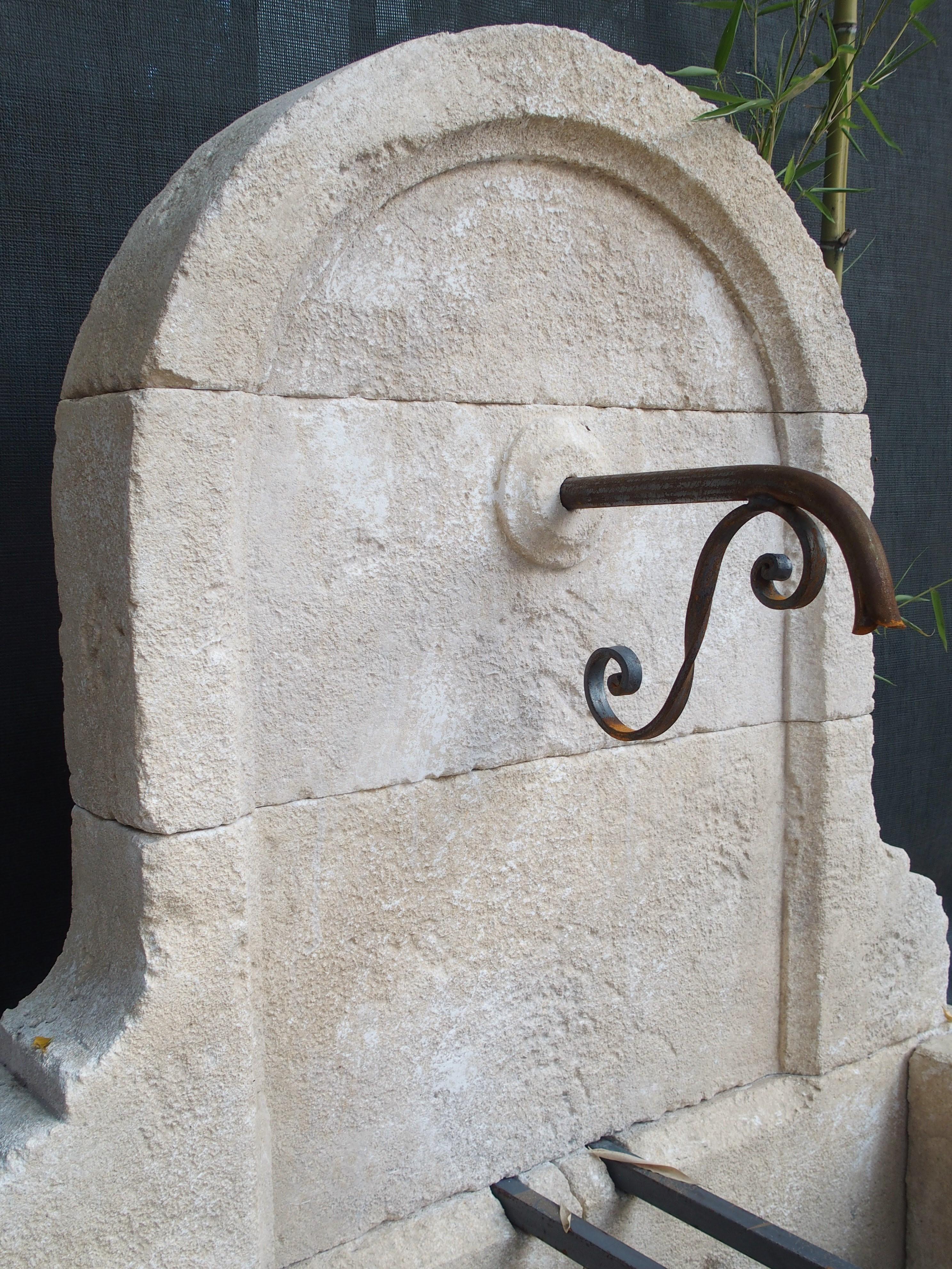 From Provence, this small arched limestone wall fountain has a curved and stepped-in molded back. It has a cast iron spout with an iron S-scroll, and the basin is rectangular with a galbe, or slightly out-curved center block at the front. Two iron