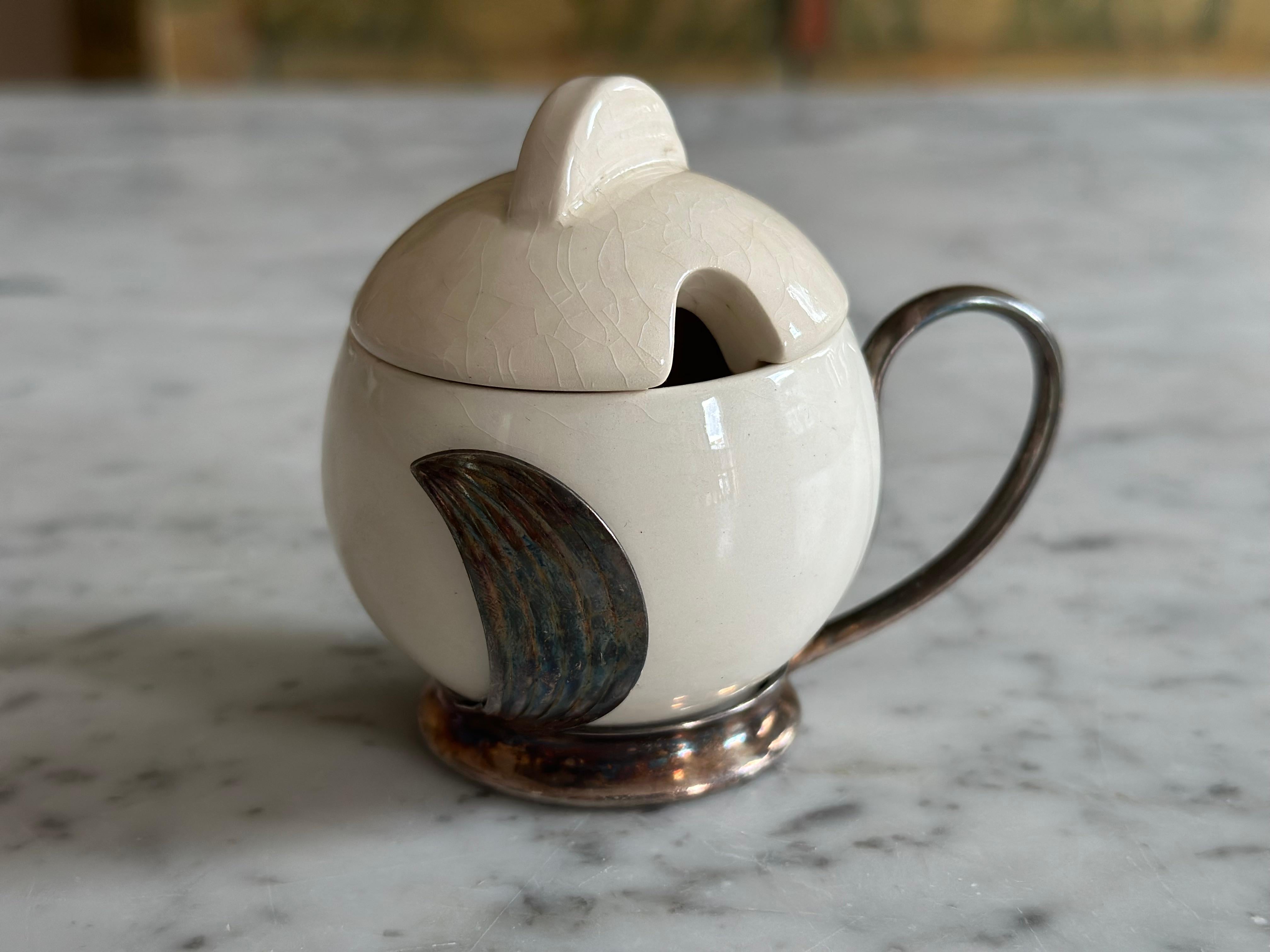 Dieser charmante kleine Senftopf ist ein schönes Beispiel für Art-Déco-Geschirr, das elegante Materialien und Handwerkskunst von zwei renommierten deutschen Herstellern vereint: WMF (Württembergische Metallwarenfabrik) und Wächtersbach Keramik. Das