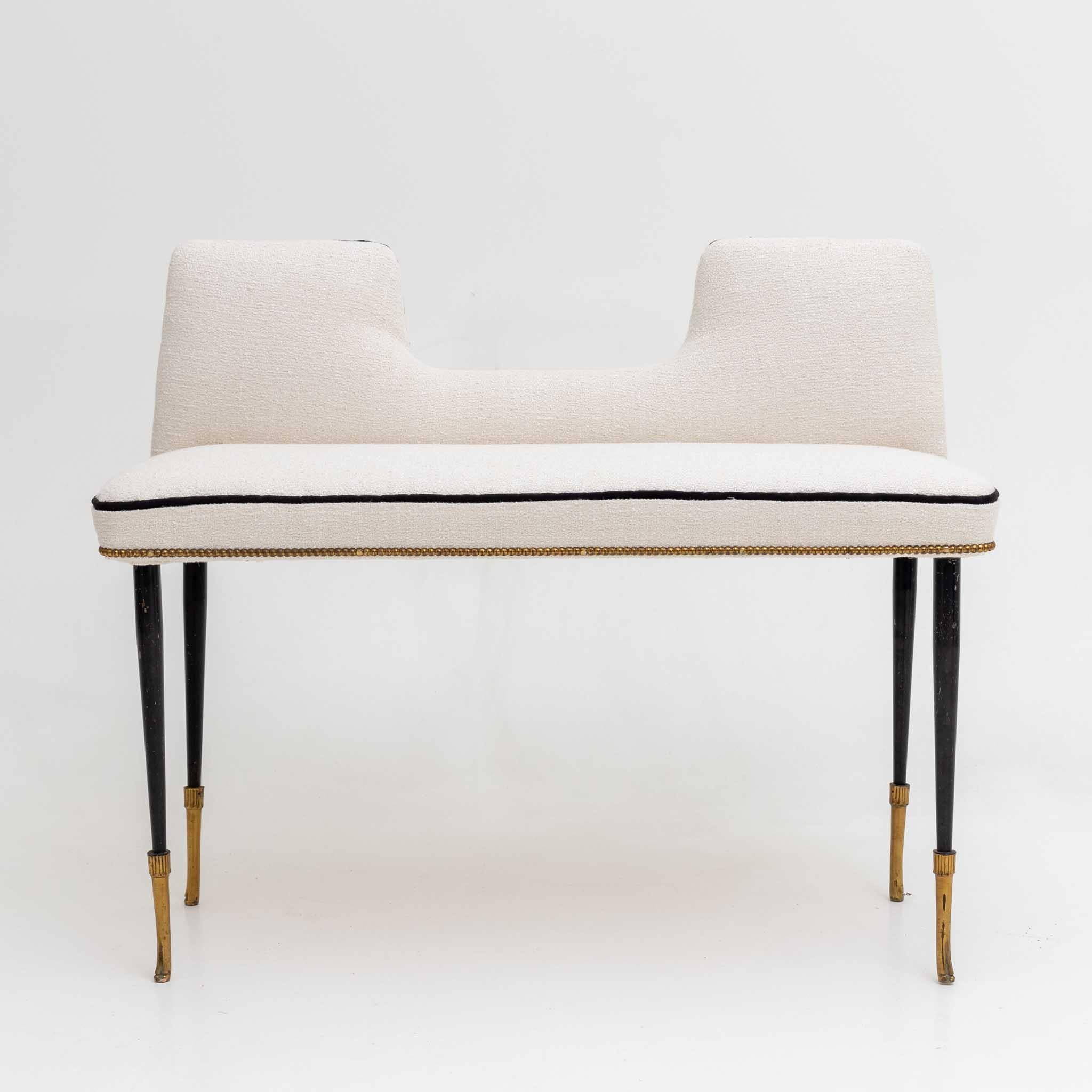 Small bench on conical black metal legs with brass sabots and seat with small backrest. The bench has been reupholstered in a white bouclé fabric with black piping and rivet decoration.
