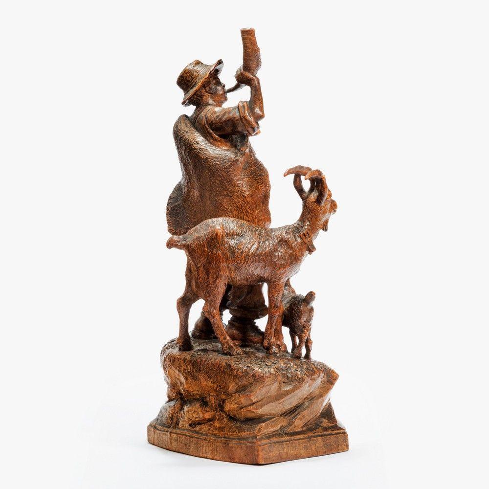 A small ‘Black Forest’ wood carving of a shepherd 
wearing a sheepskin cloak and blowing a Horn, with mountain goats at his feet, stamped SR with a paper label, indistinct ‘Brugger, Interlaken’.