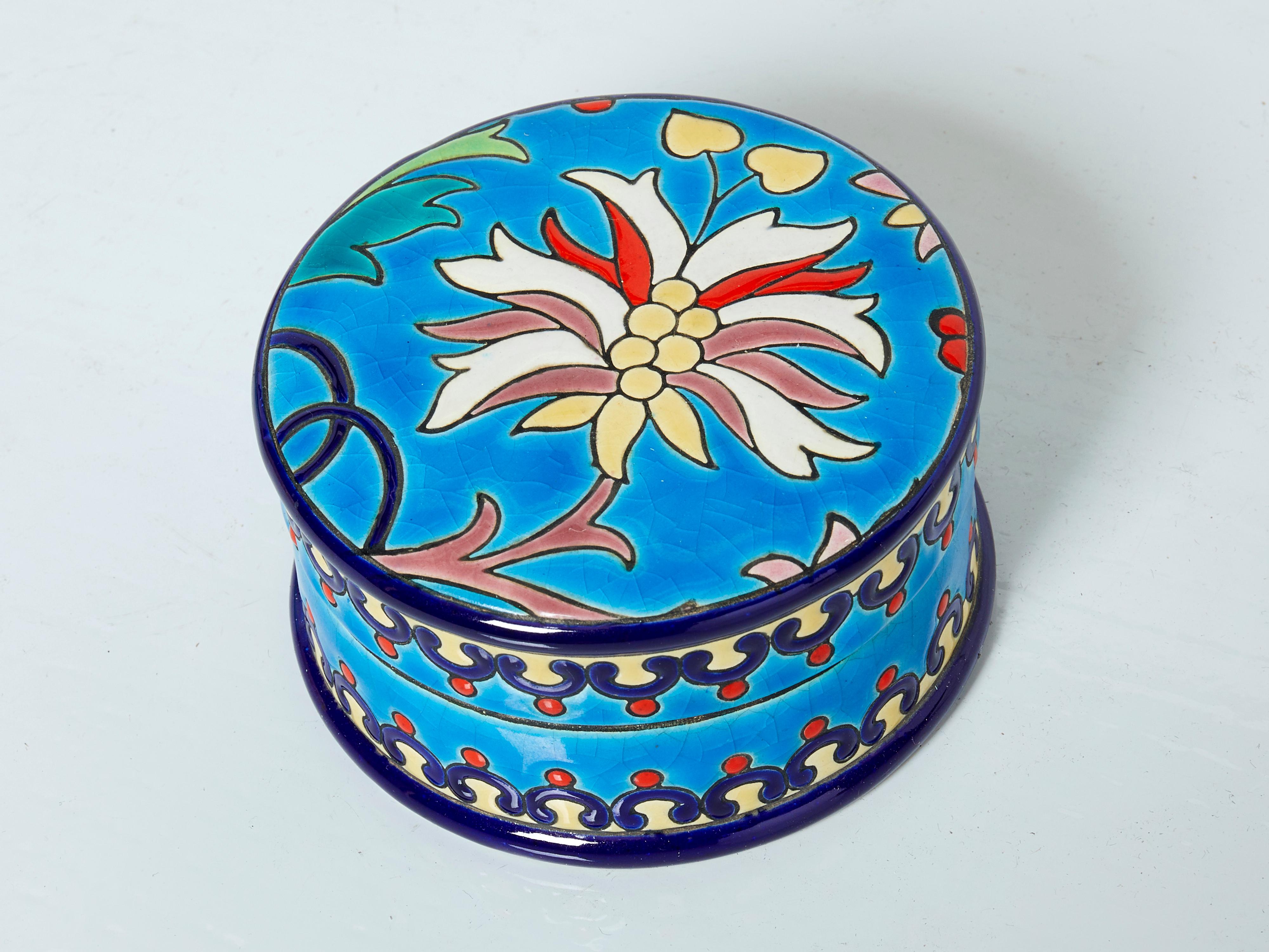 Bunte kleine runde Keramikdose im Art déco-Stil von Faïenceries et Emaux de Longwy aus der Zeit um 1940. Diese türkisblaue Dose hat einen farbenfrohen Fries mit einer schönen Blume auf der Oberseite und die für Longwy typische Craquelé-Glasur. Die