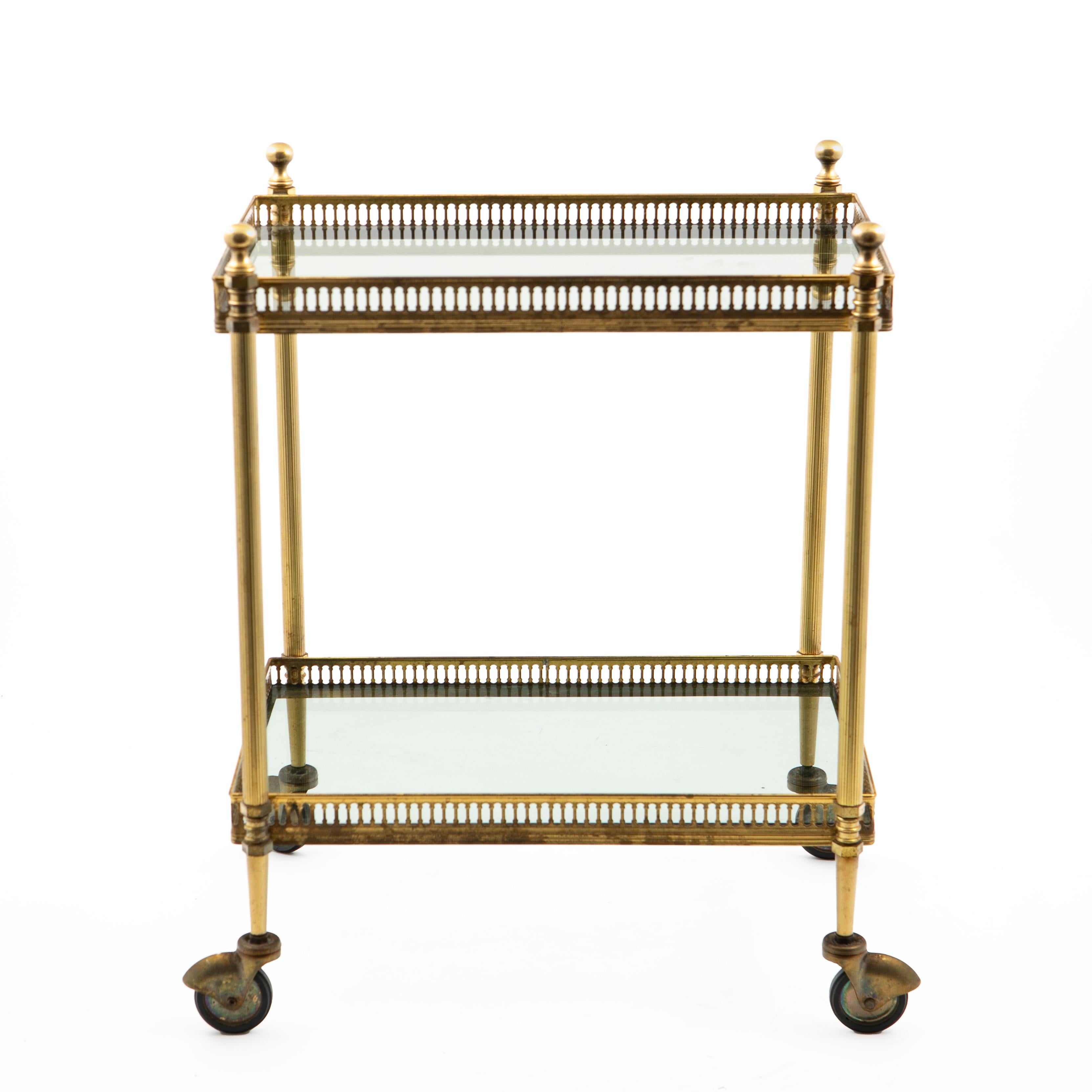 Small two-tiered brass serving trolly or bar cart featuring two smoked glass shelves surrounded by pierced brass galleries.
Round and fluted legs with decorative finials endning in castor wheels.
Original good vintage condition, nicely