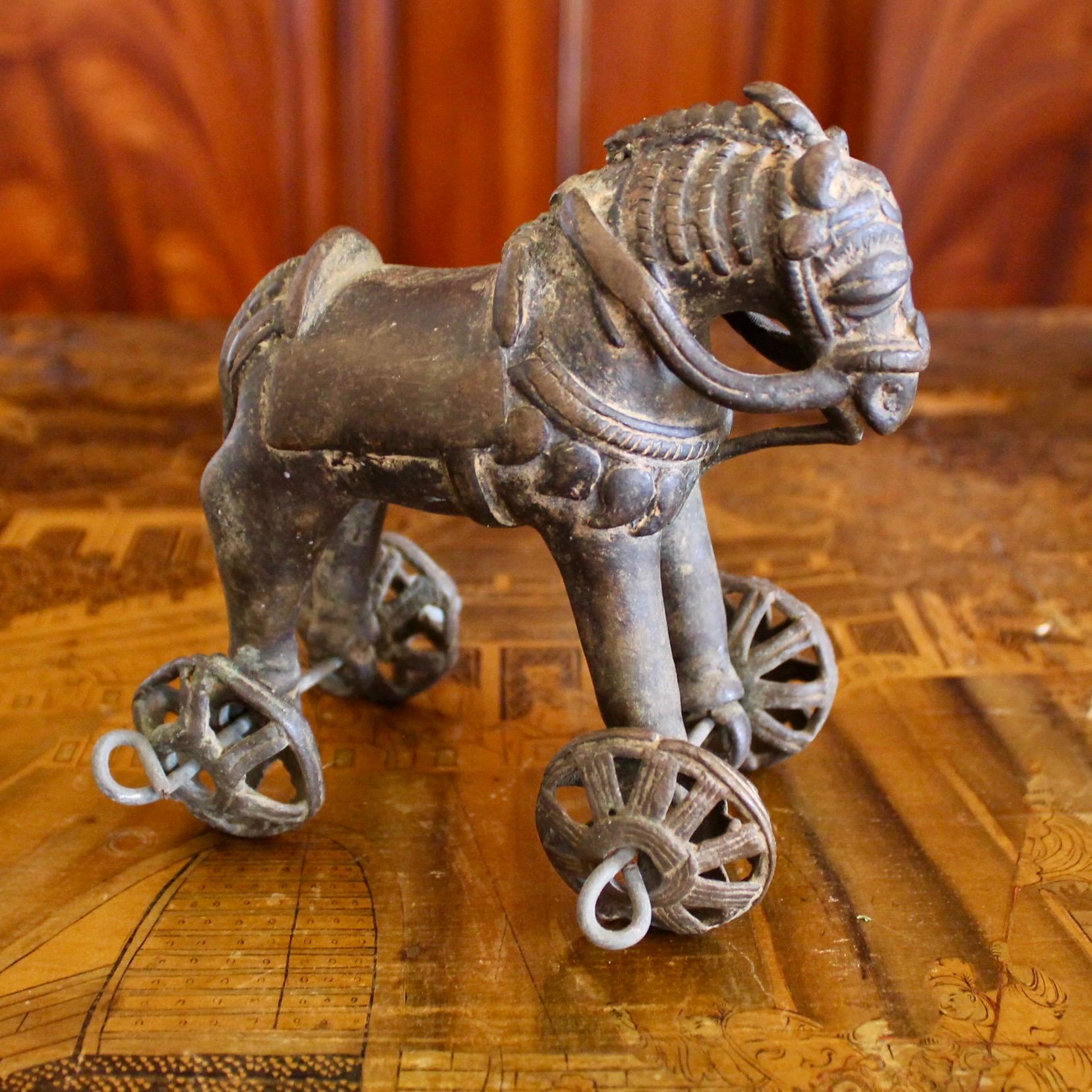 A charming cast bronze horse with elaborate caparisoned high pommeled saddle and trappings, and a braided mane and tail, the legs ending in wheeled axels. This was a temple offering, of a type known as “temple toys,” left by the devout at Hindu
