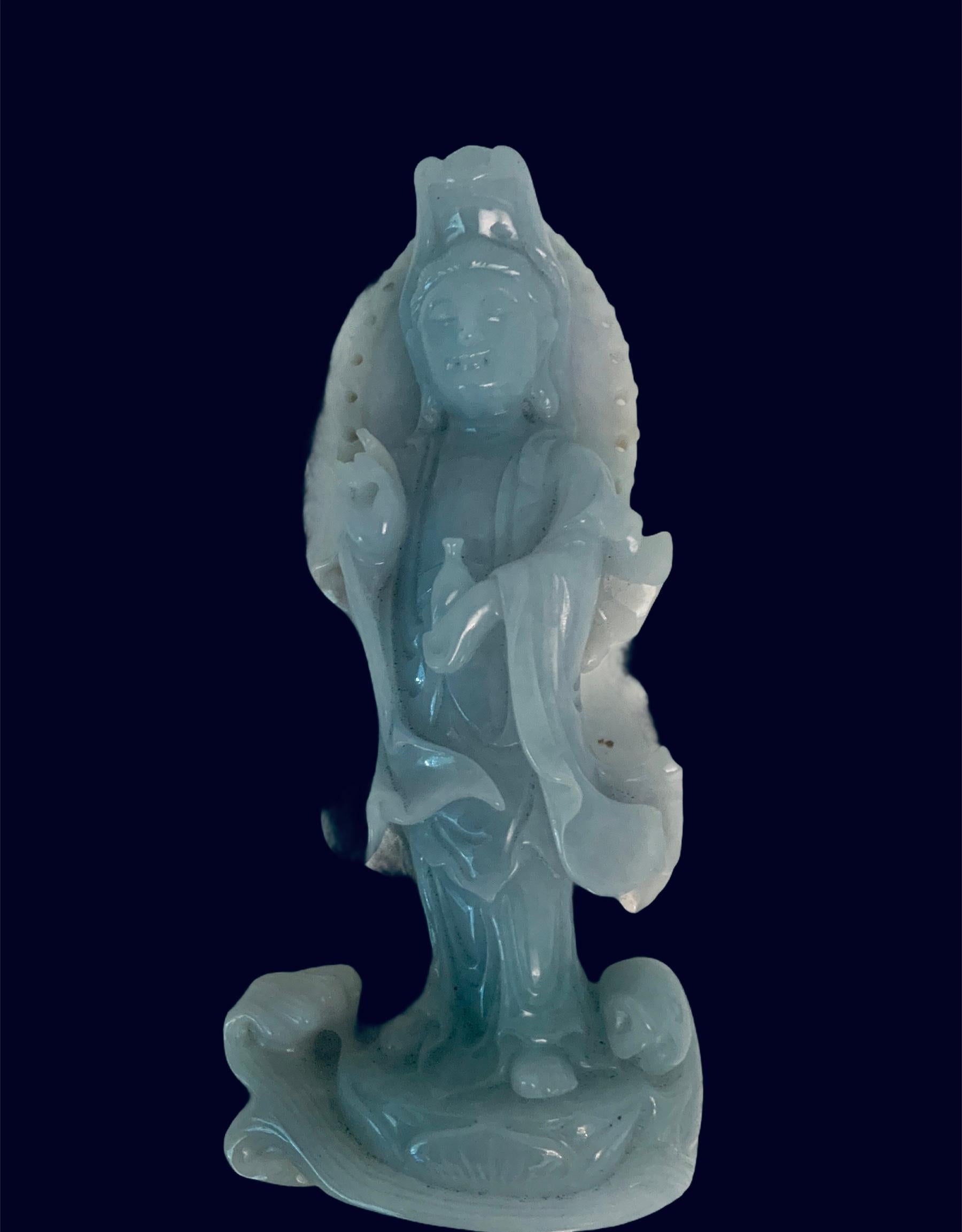 This is a small carved lavender “opaline” jade Guan Yin sculpture. The sculpture has some russet jade spots in the upper back. The back of her head is adorned with a reticulated mandorla. She is dressed with a lively buoyant robes. She is holding a
