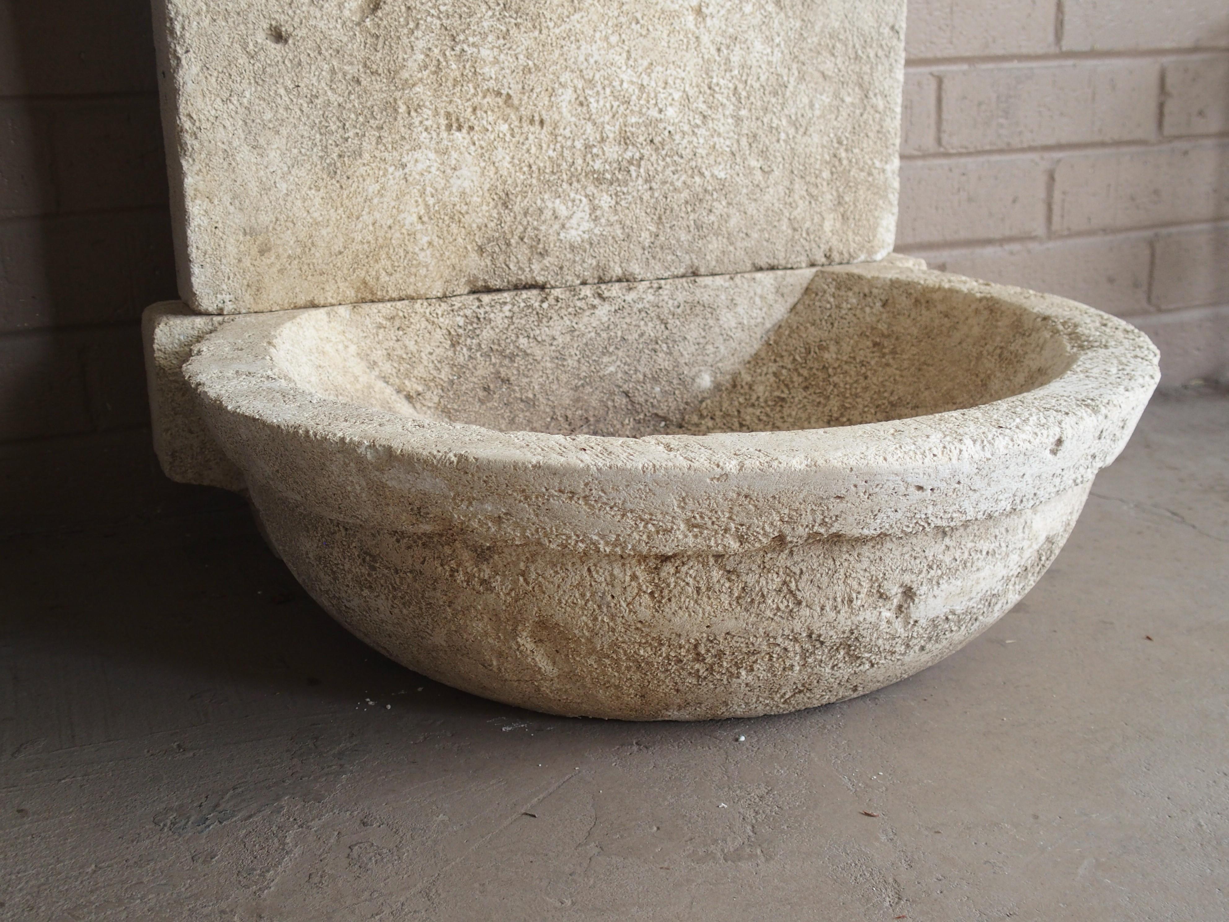 This small hand carved limestone fountain from France has a circular basin with hole for drainage. The basin extends out from a vertical back support, and on top of this, is a carved upper portion with sloped shoulders. The rusted spout slots into a