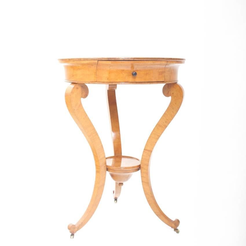 Small table with one drawer on three legs, with a circular tabletop out of ash tree. The tabletop shows a beautiful floral inlay with a framing meander motif.