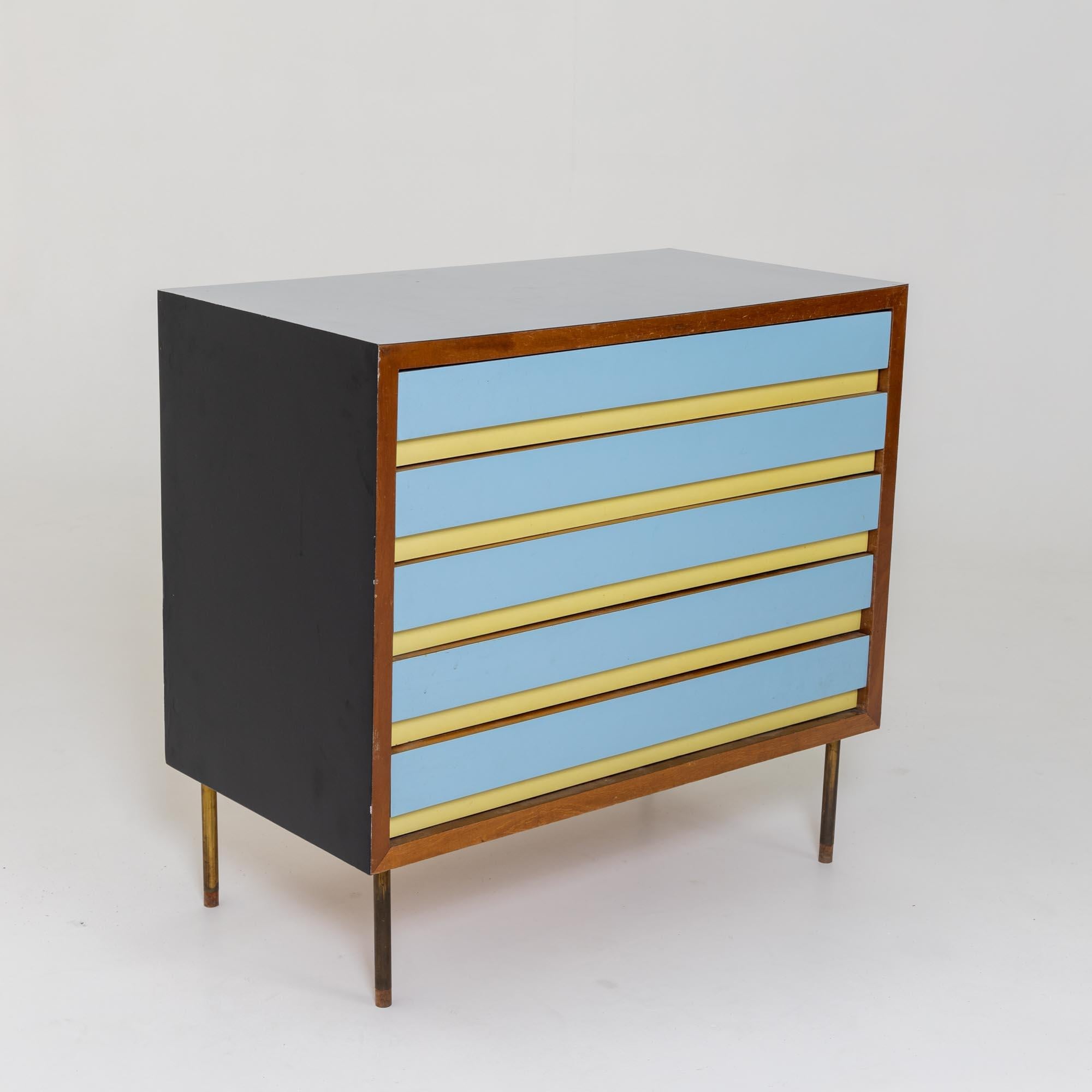 Small chest of drawers with five drawers on conical brass legs. The surfaces are light blue and the fronts of the drawers are set off in light blue and yellow.