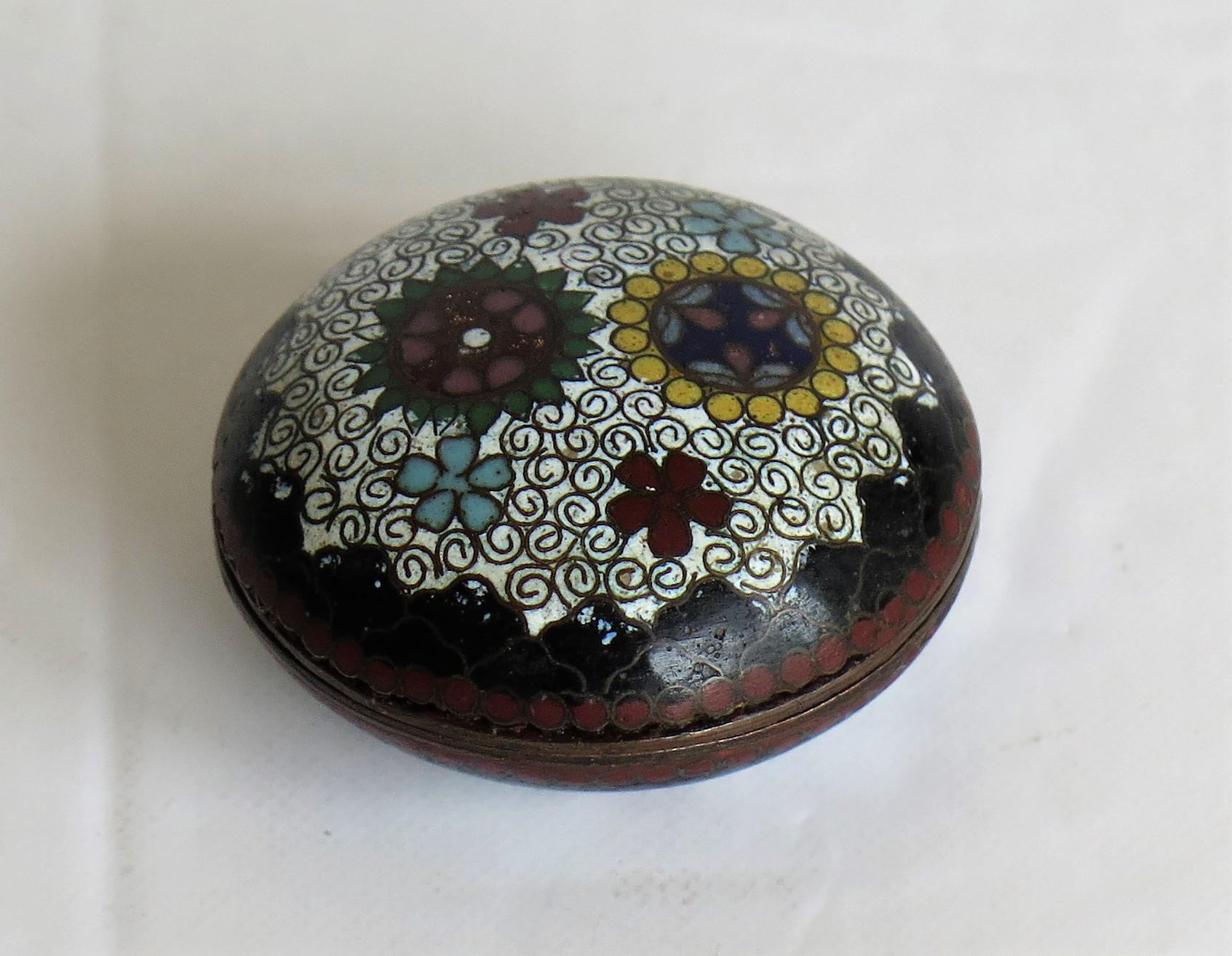 This is a very decorative small cloisonné lidded box, made in China during the Qing dynasty and dating to the mid-19th century.

The box has a circular shape with a domed lid. It has been well made of a bronze alloy with enamels of many different