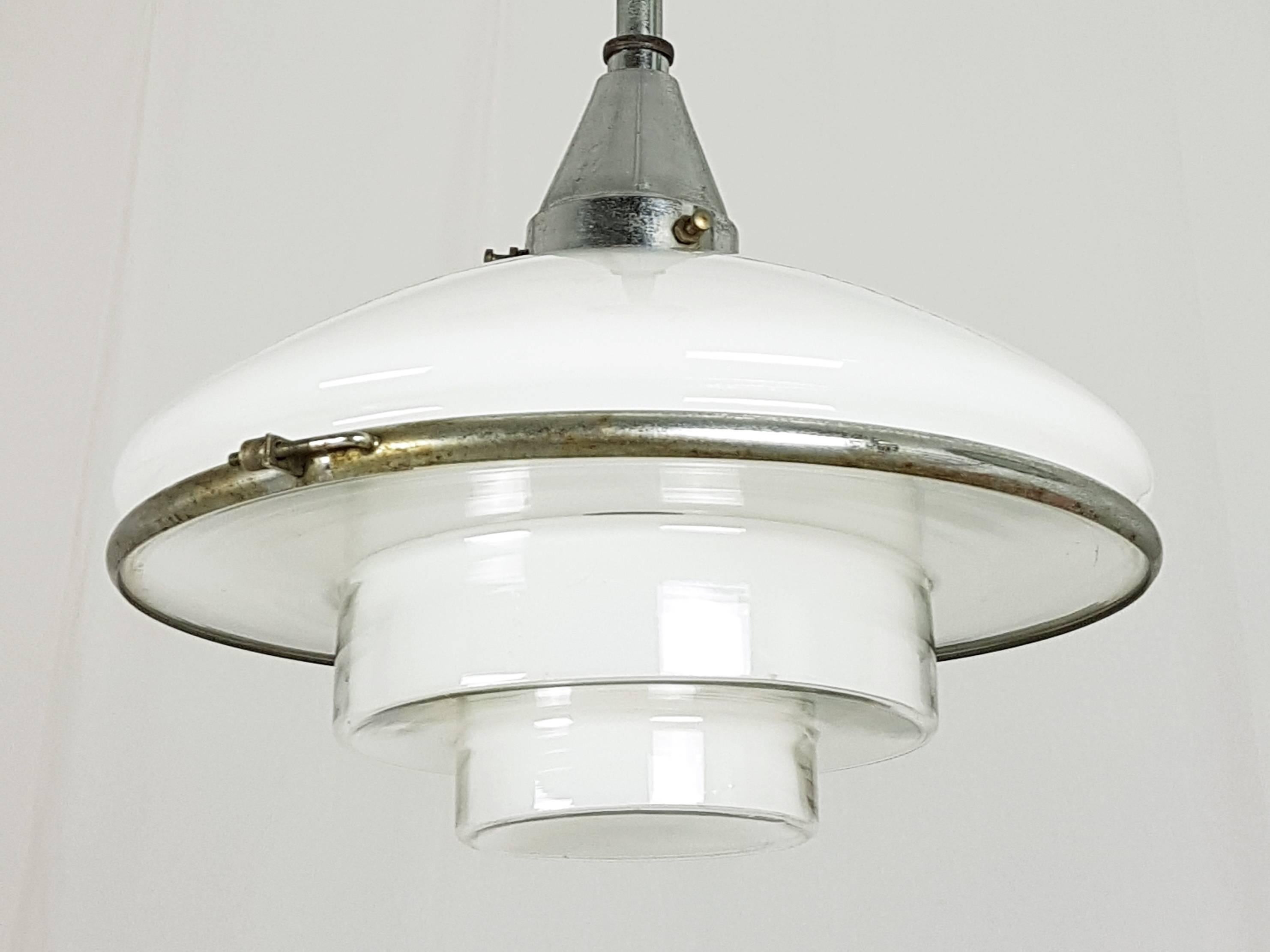 This pendant lamp was designed by Otto Müller in 1931 and manufactured by Megaphos/Sistrah Licht GmbH in Germany. It is made from 2 glass shades joint together by a chrome-plated belt. The upper shade is made of white opaline glass; the under shade