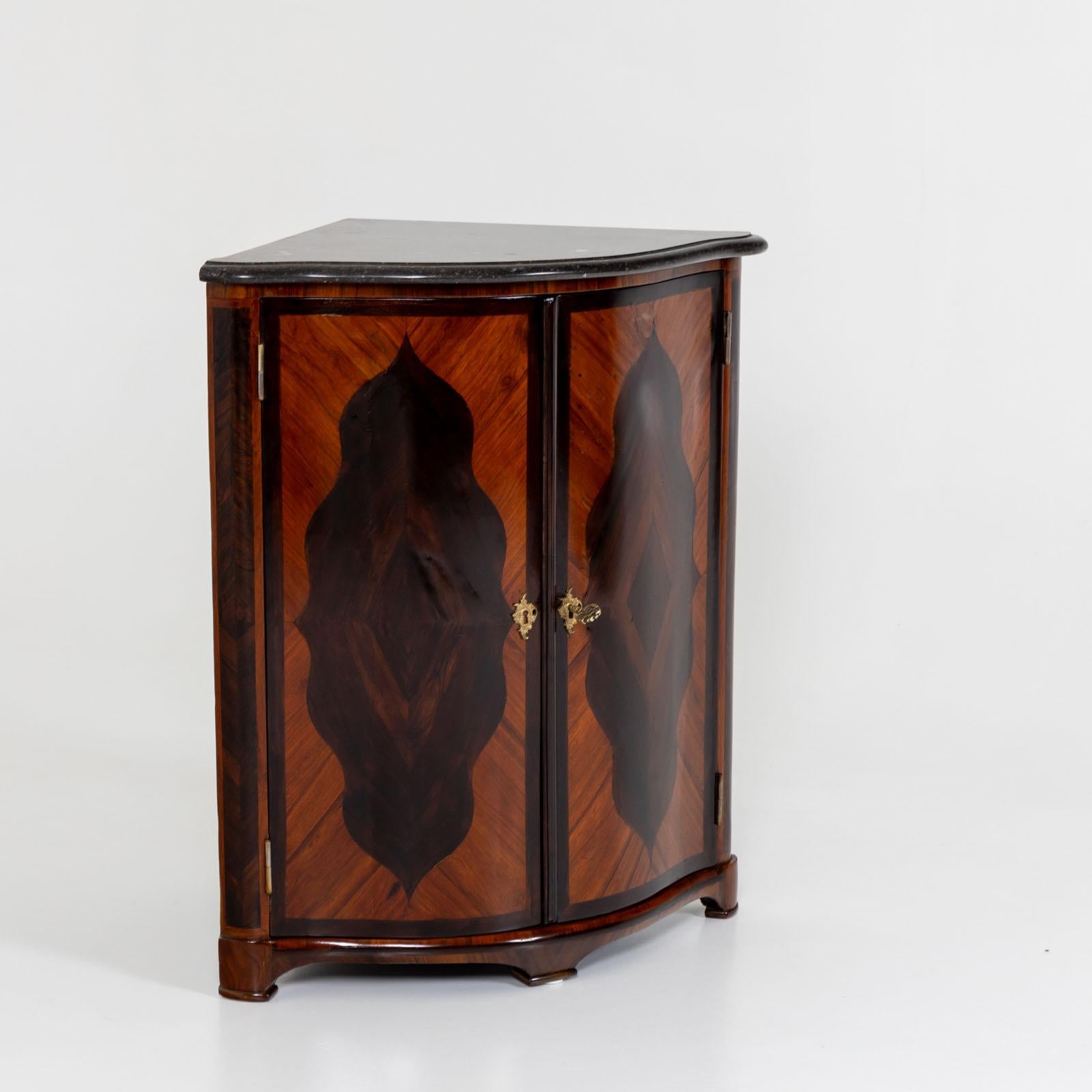 Pequeño armario esquinero de dos puertas con encimera de piedra oscura moldeada y frente redondeado. Las puertas llevan incrustaciones de madera de colores contrastados, formando una elegante cartela de múltiples curvas. El interior está dividido en