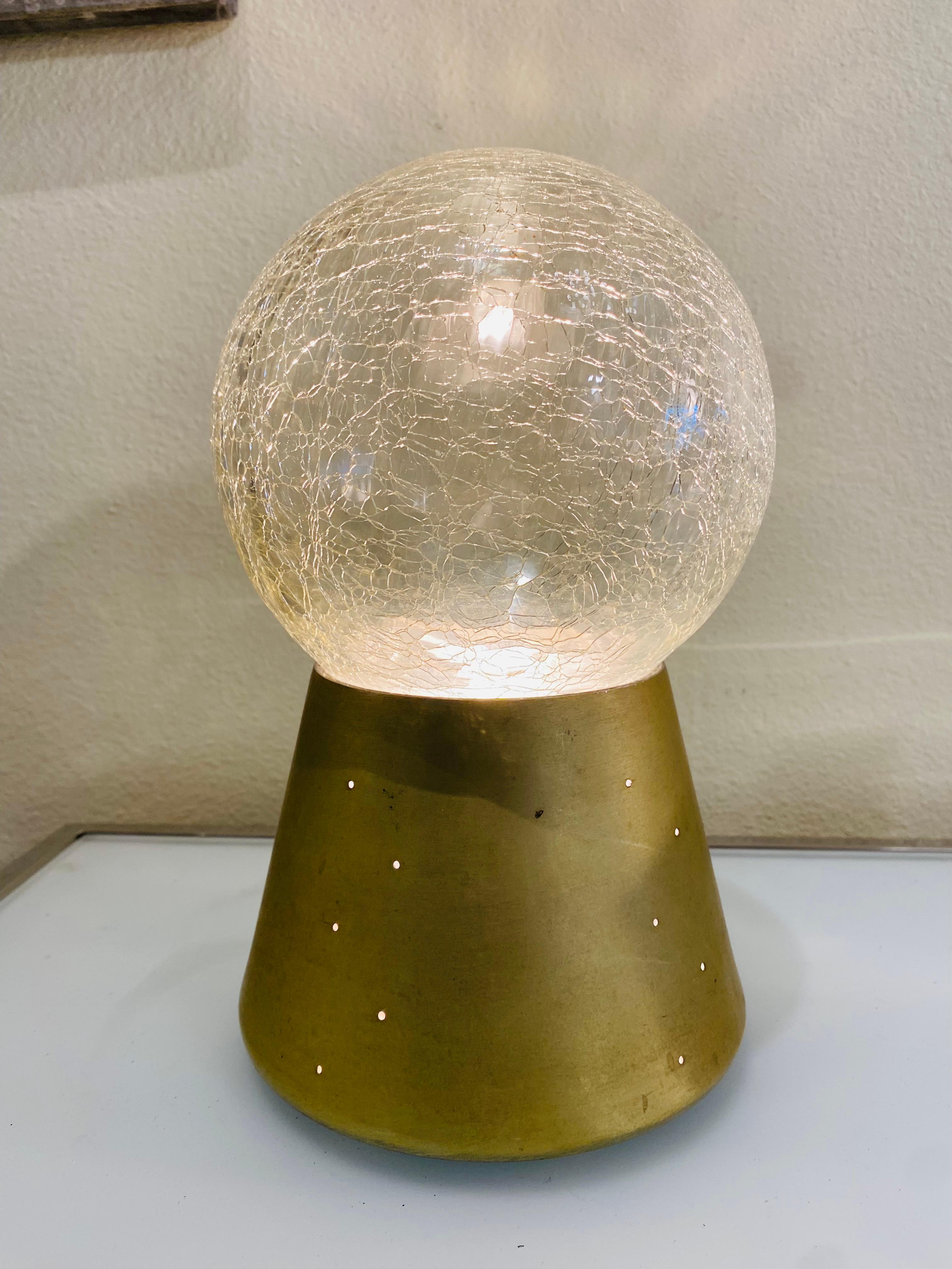 This is a table lamp featuring a crackled glass globe resting on a conical gilded aluminum base. The base has small perforations that allow light to diffuse subtly, complementing the soft glow from the glass sphere above.