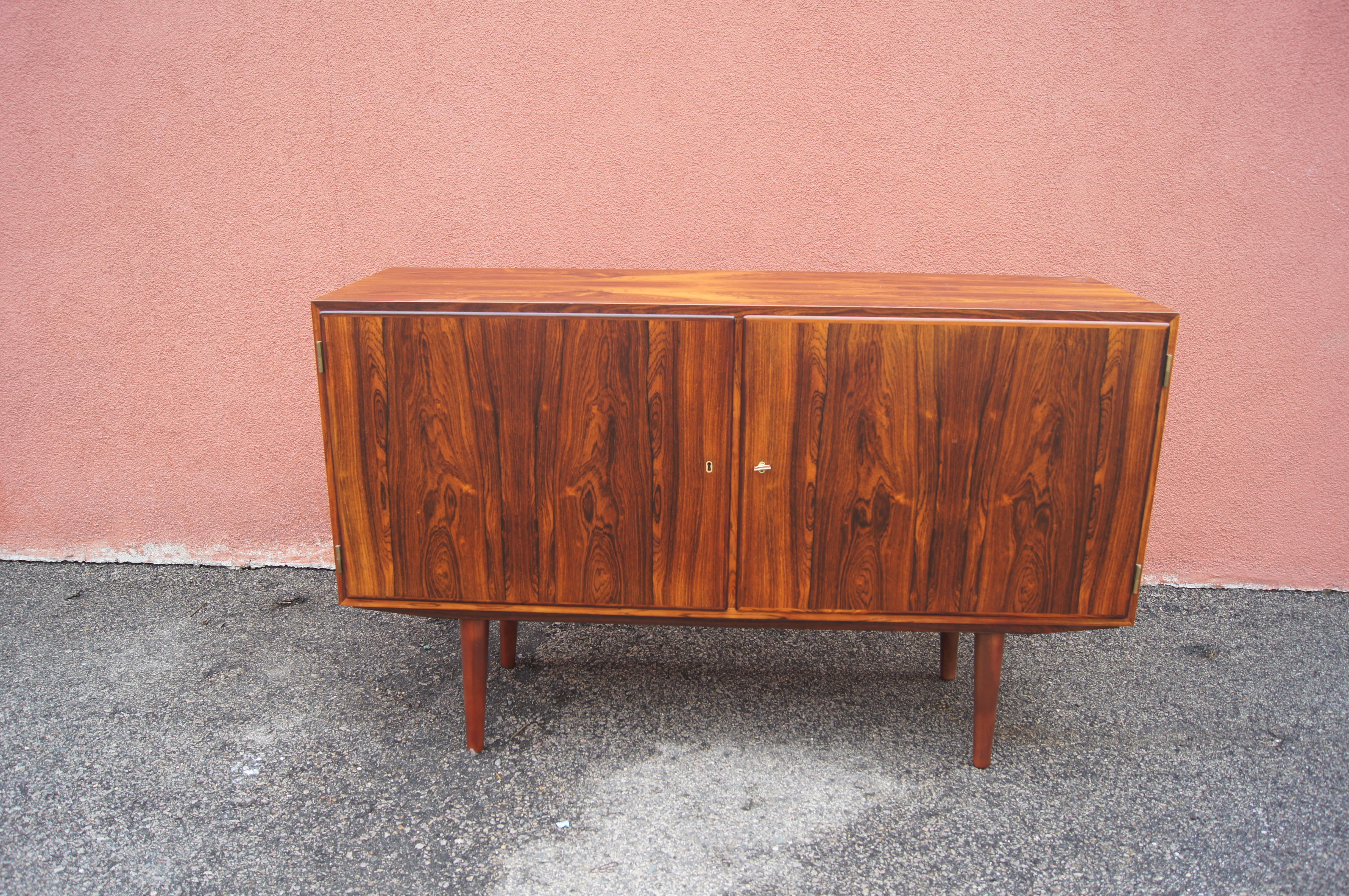 This Danish modern credenza comprises a small rosewood case on solid rosewood legs. Behind the two locking doors are three adjustable shelves on the right and four shallower felt-lined tray shelves on the left.

The two locks share the original key.