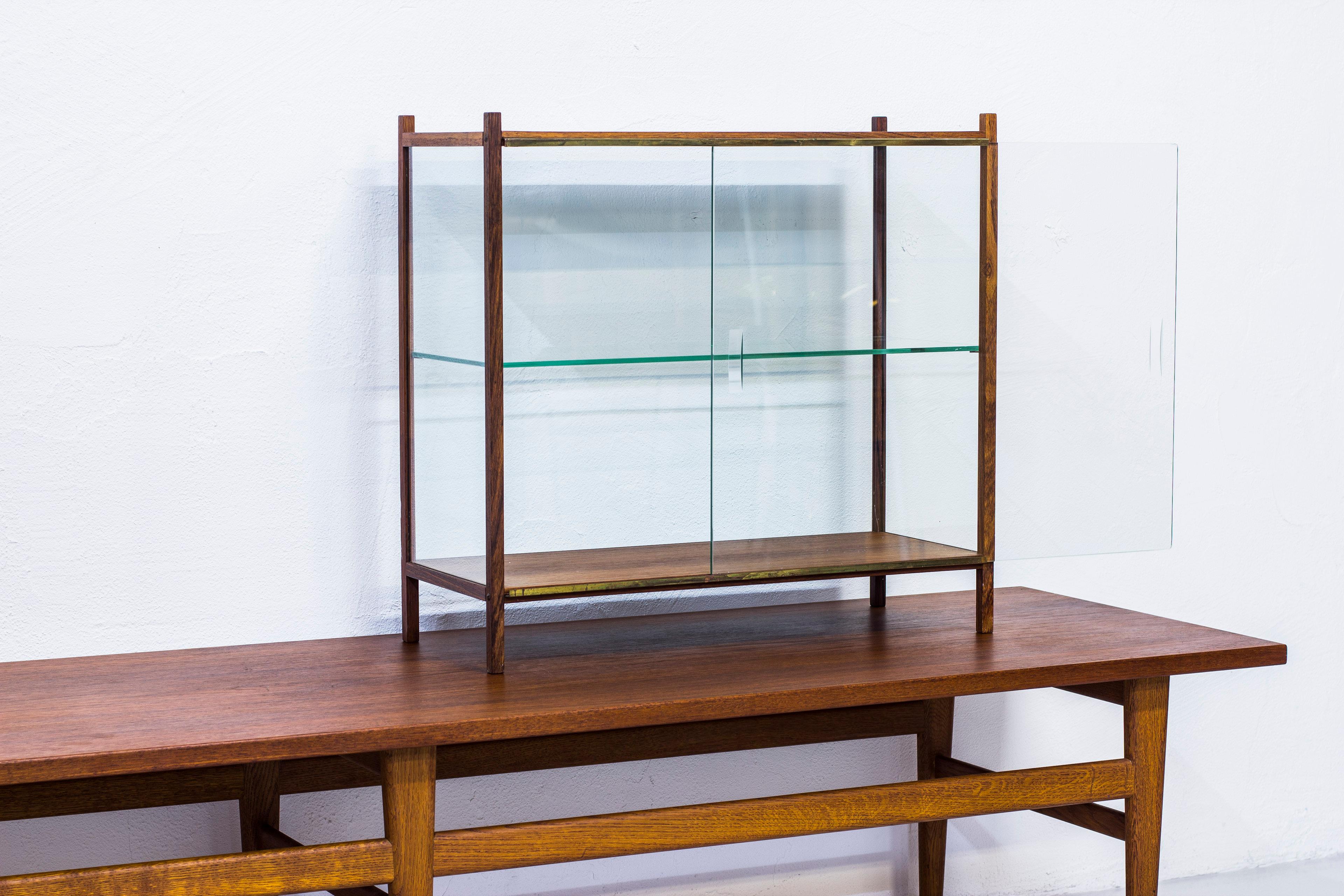 Small display cabinet or vitrine made in Sweden during the 1960s. Made from rosewood, glass and brass. Very good vintage condition with minor signs of usage and patina. Sliding door of glass with cut out handles. One optional glass shelf on the