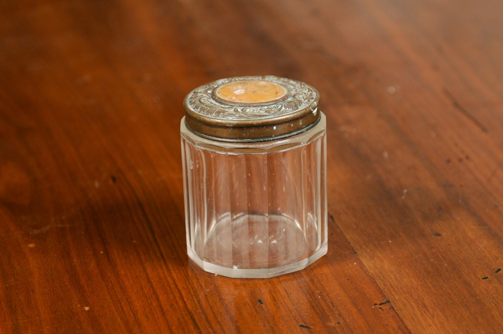 A small English Victorian period glass vanity jar from the 19th century, with metal lid and faceted design. Created in England during the 19th century, this jar container features a circular glass body adorned with faceted patterns. Topped with a