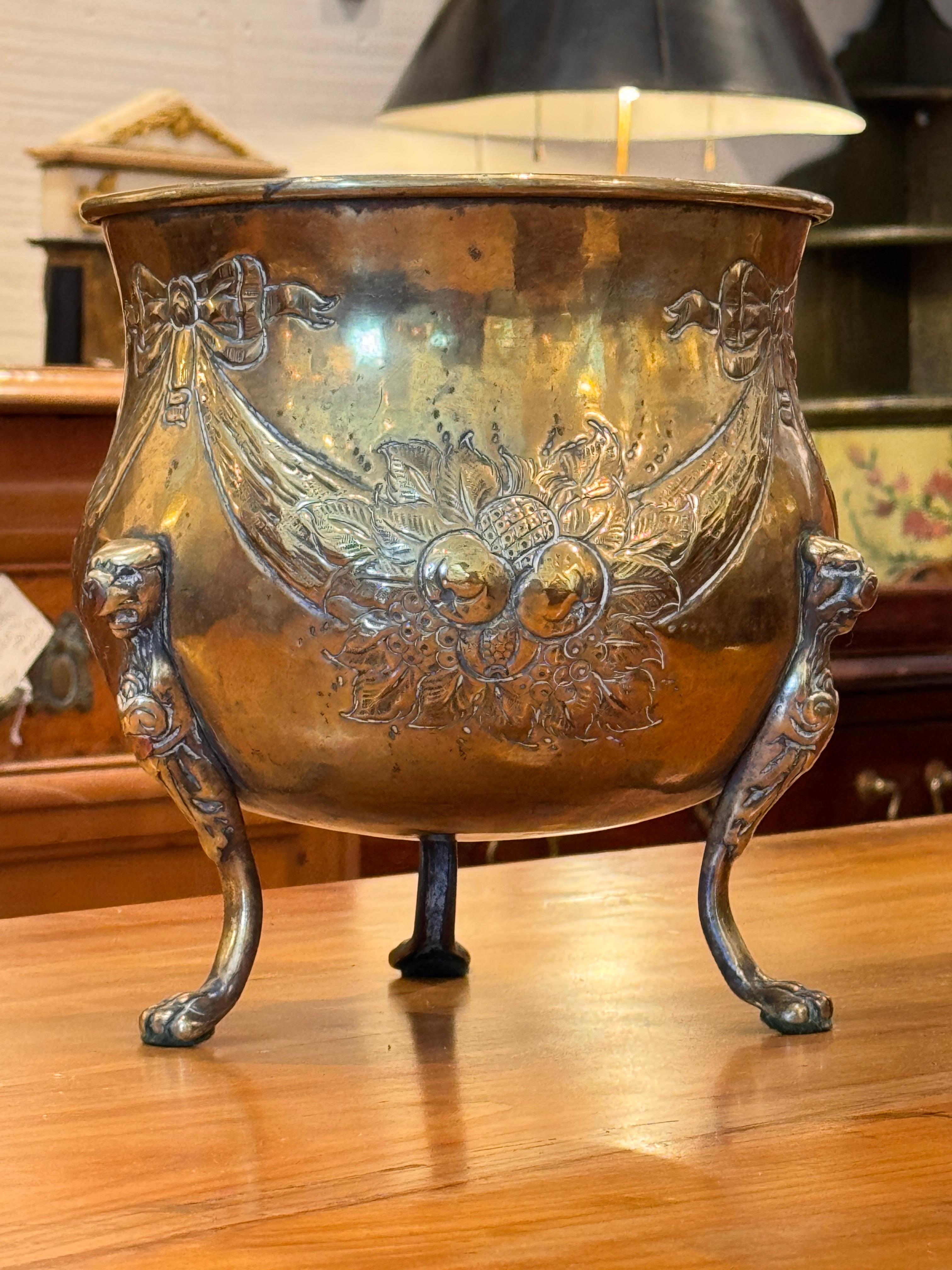 A small brass planter raised on three paw feet, with embossed decoration featuring ribbon-tied swags and a central fruit and foliate motif. The warm patina and elegant detailing give this piece both charm and presence