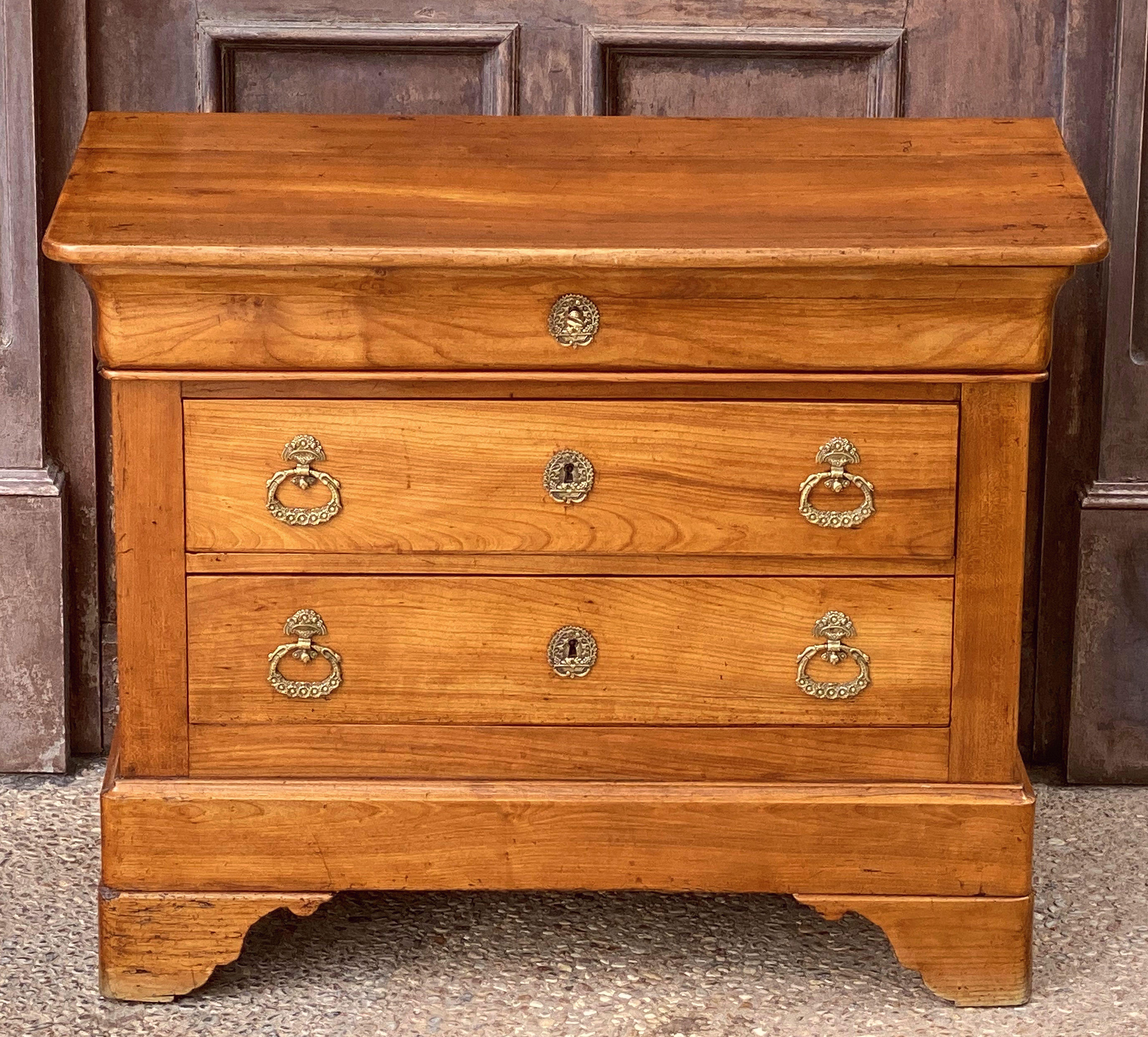A fine French small chest of drawers or commode of patinated cherry, featuring a moulded top over a paneled base with three drawers, with original brass handles and escutcheons, with key, and resting on bracket feet.
  