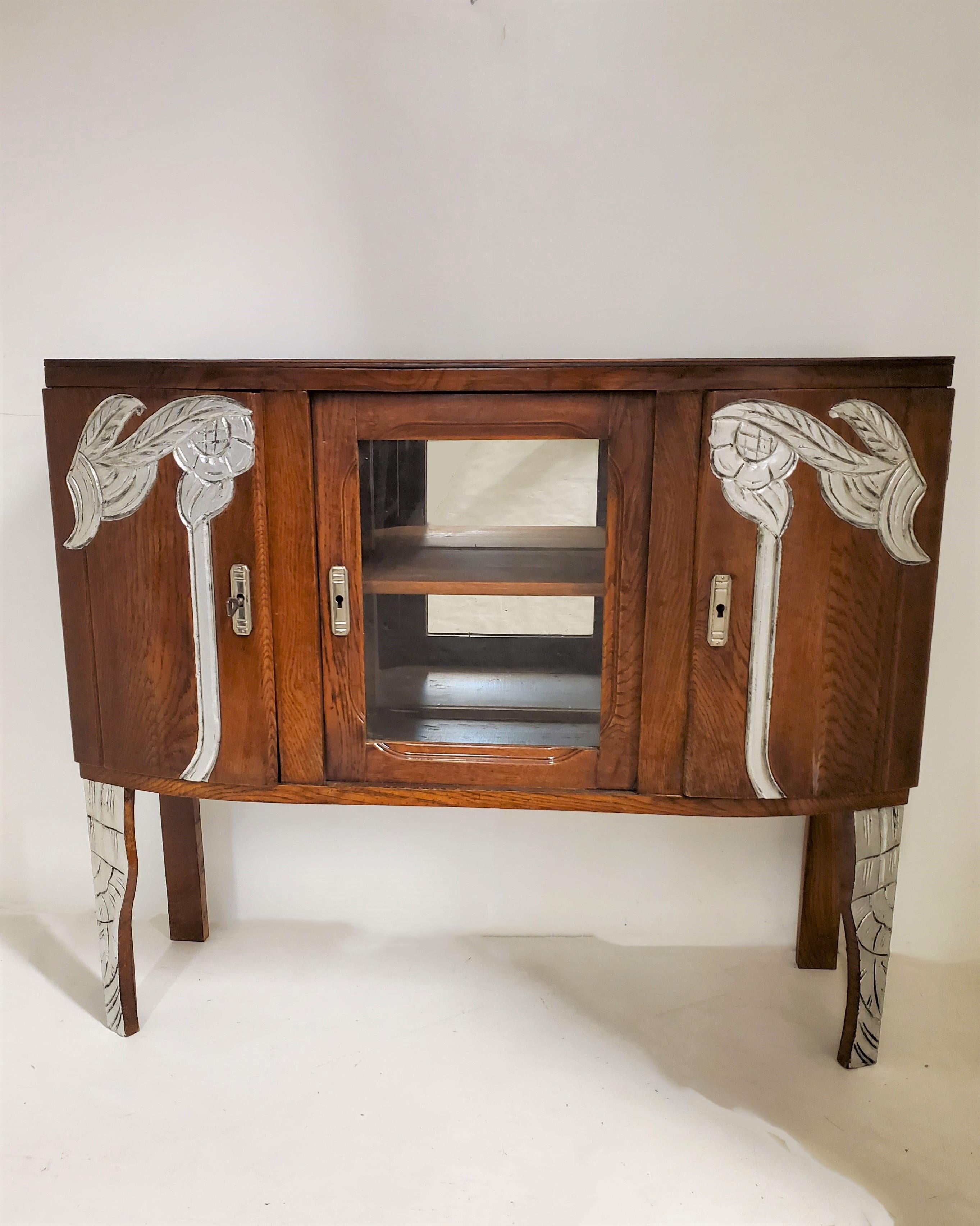A petite French Art Deco three door European oak cabinet with hand carved metal leaf detail of stylized floral, leafage and linear design. 
The center of the cabinet houses a mirrored backed glass display case.
Geometric, fluted and stylized