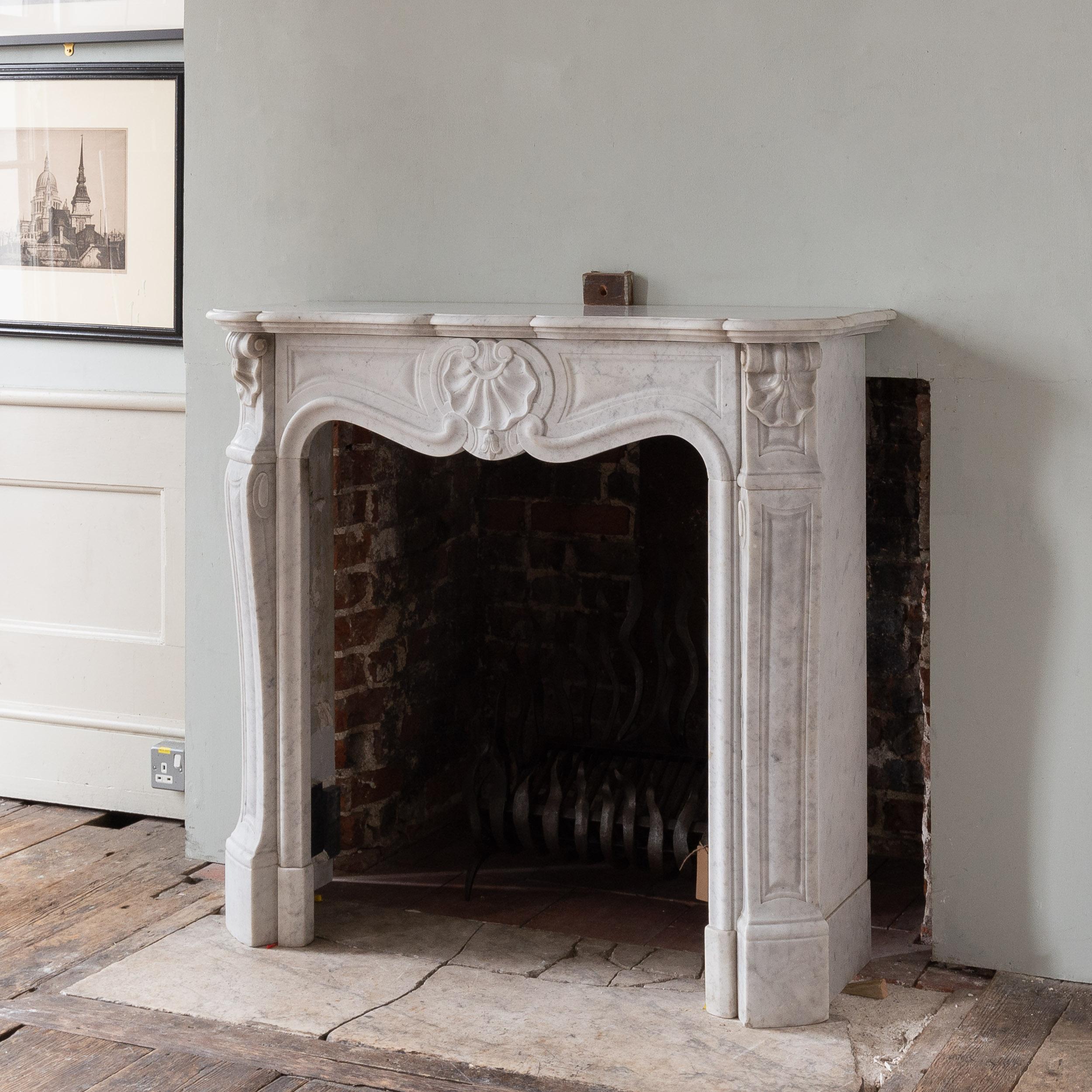 A Small French Rococo Carrara Marble fireplace, dating to the early twentieth century in the Louis XV style, with scallop shell to the centre of the shaped frieze and panelled frieze, the out-turned volute jambs with scallop shelf clasps supporting