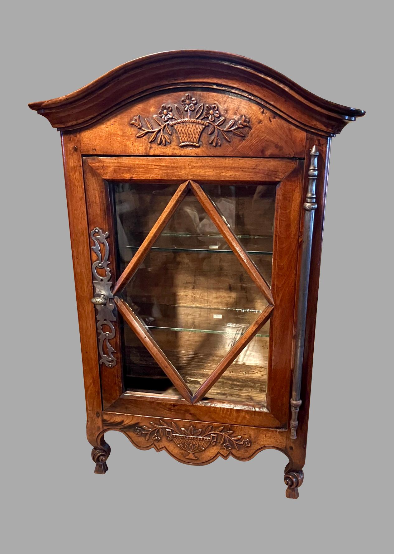 A lovely walnut French provincial cabinet of small size, the arched moulded cornice with a charming carved design of potted flowers above a paneled glazed door with with a polished steel escutcheon resting on upturned scroll legs. The apron is