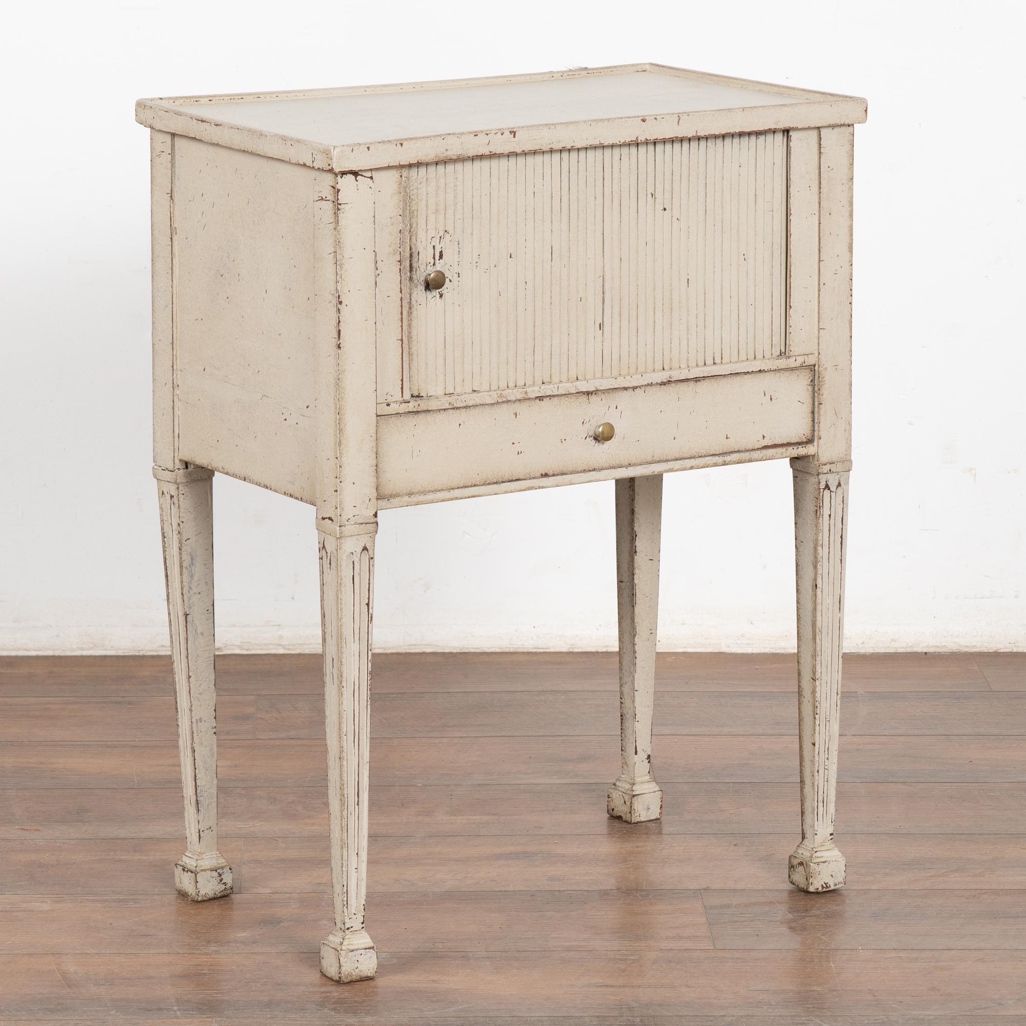 Small Swedish country gray painted cabinet standing on fluted tapered legs; may serve also as a nightstand or side table.
A single fluted drawer with brass pull rests under the cabinet which opens with a sliding pocket door.
The light dove gray