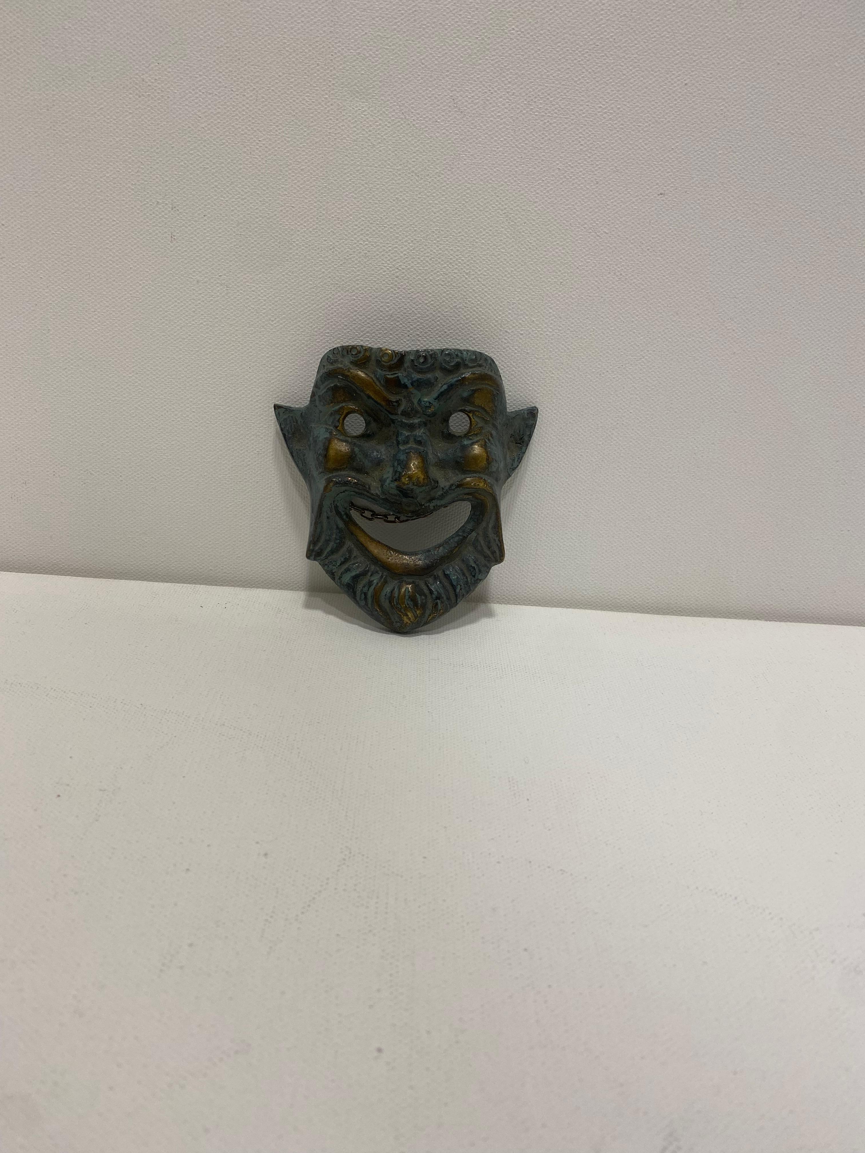 Dies ist eine kleine griechische Theatermaske aus Bronze aus den 1970er Jahren, die für die Wandmontage bestimmt ist.

Die Maske hat einen übertriebenen Gesichtsausdruck, der typisch für die antiken griechischen Theatermasken ist, die bei