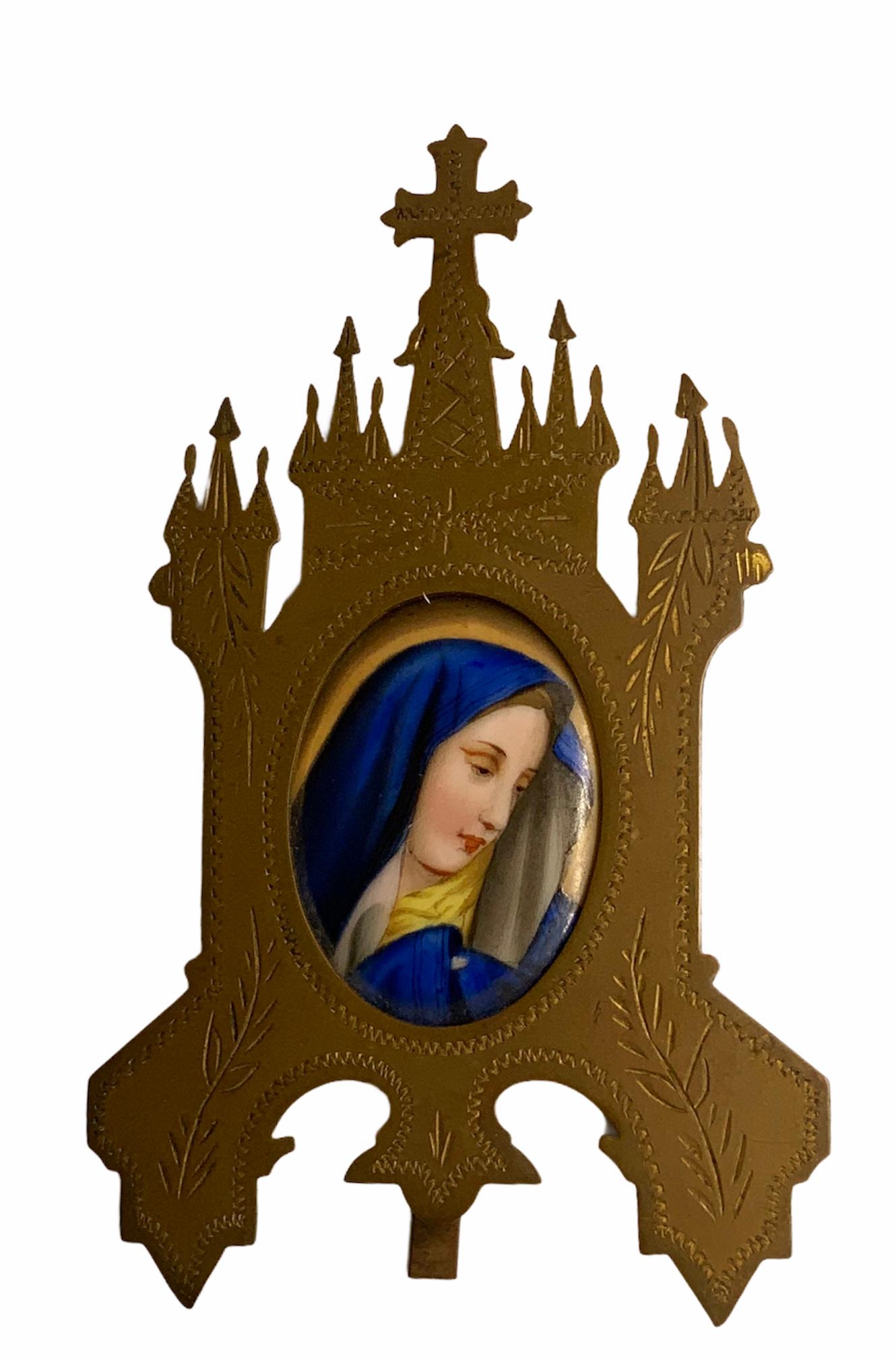 This is a hand painted porcelain oval portrait plaque of the Virgin Mary for home devotion. It is framed in a golden brass metal shaped as a gothic church that serves as a stand. The brass is adorned with some carvings of foliages and zigzags