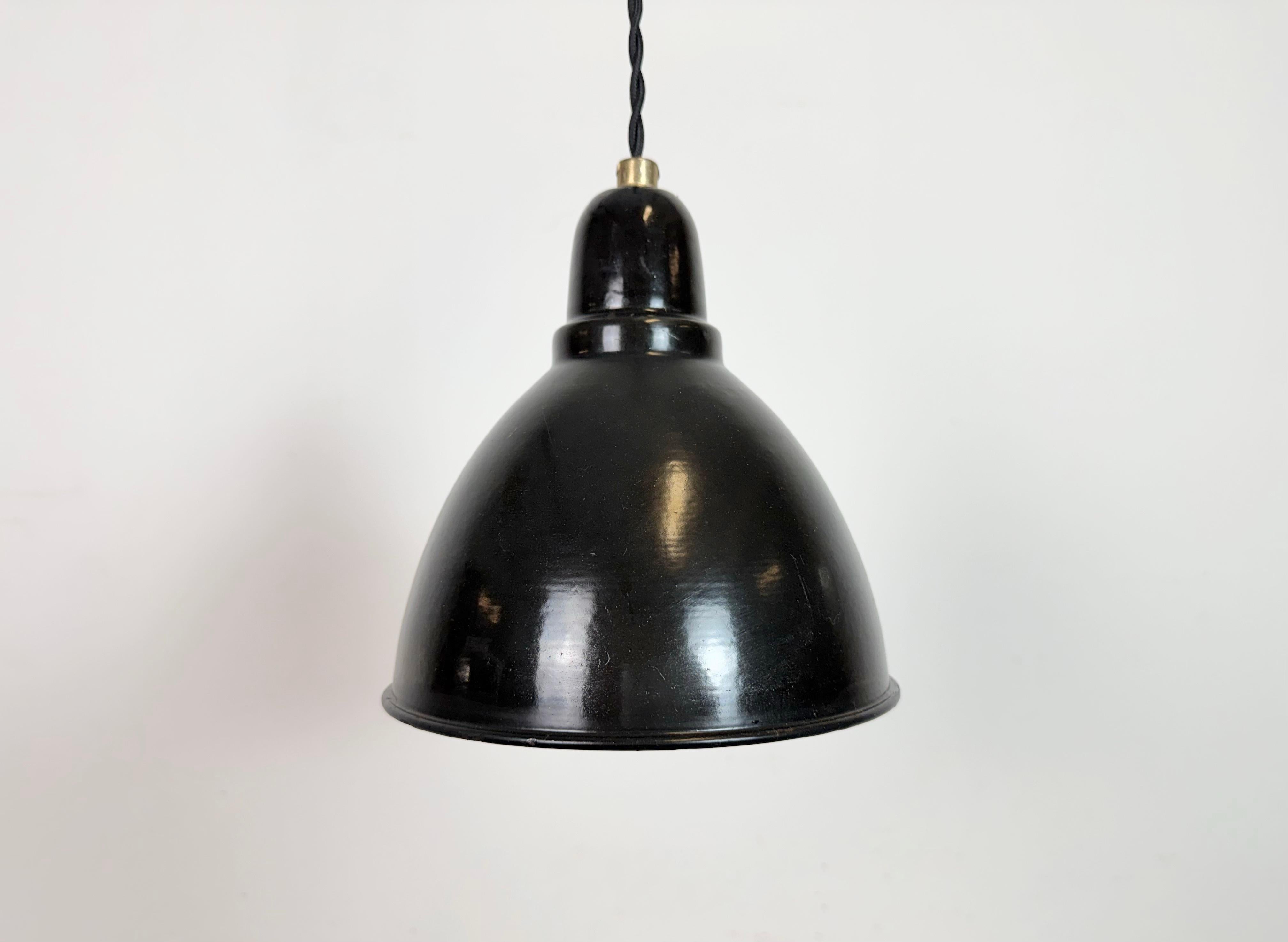 Lámpara colgante industrial de esmalte negro fabricada en la antigua Checoslovaquia en la década de 1950. Esmalte blanco en el interior de la pantalla. Marquesina de celosía metálica. El zócalo de porcelana  requiere bombillas estándar E27/ E26.
